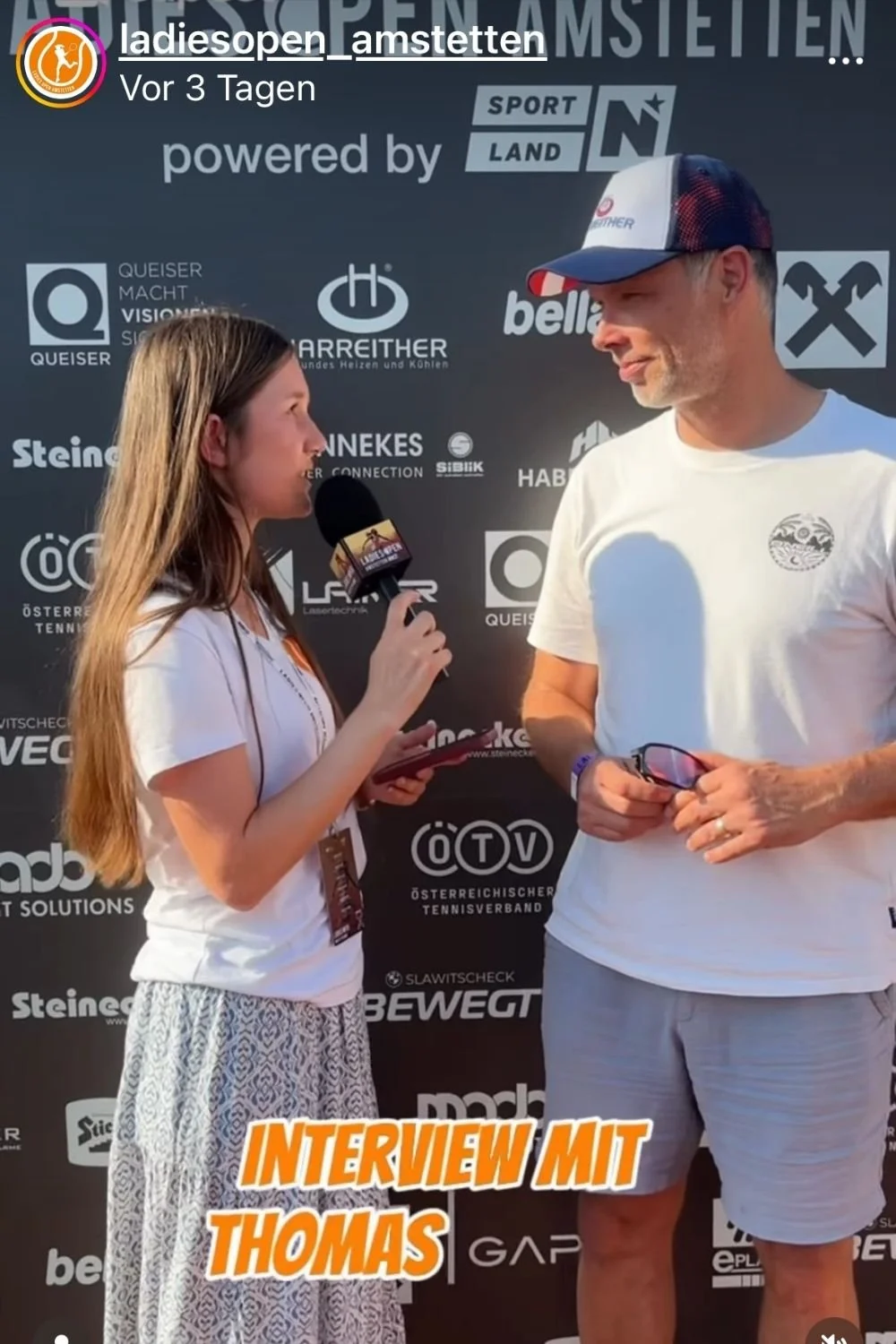 Eine junge Frau, die Mitglied des Social Media-Teams ist, interviewt Skistar Thomas Sykora Mann vor der Sponsoren-Wand beim Damen-Tennisturnier in Amstetten. Die Frau hält ein Mikrofon und einen Notizblock.