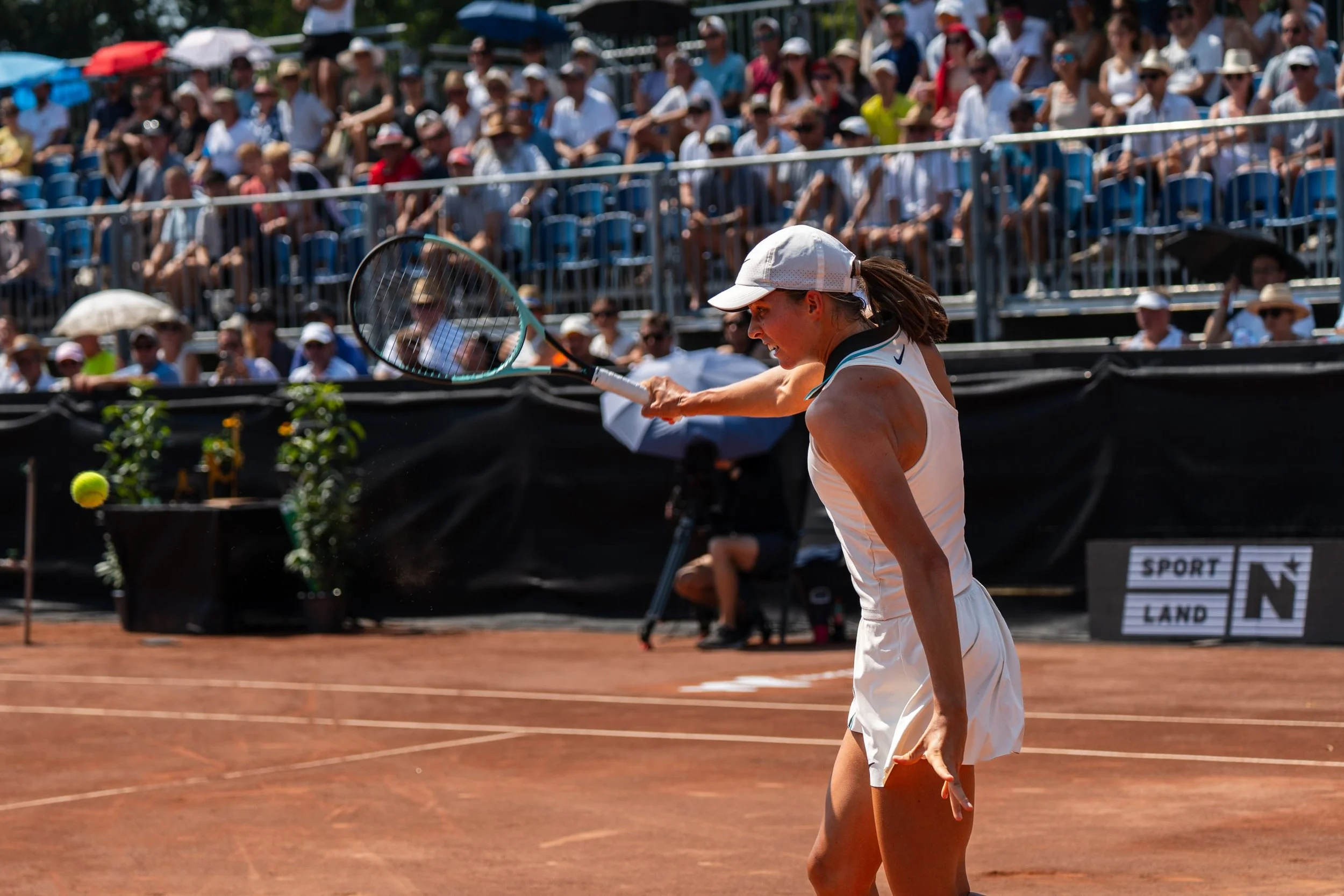Eine Tennisspielerin im weißen Kleid beim Schlag auf einem roten Tennisplatz während eines Spiels vor einem großen Publikum.