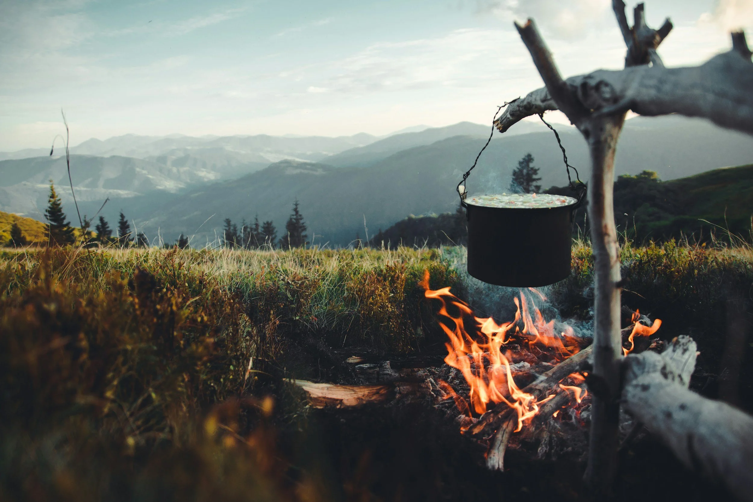 A small campfire with a black pot hanging over it, set in a mountain landscape with rolling hills and pine trees, under a partly cloudy sky.