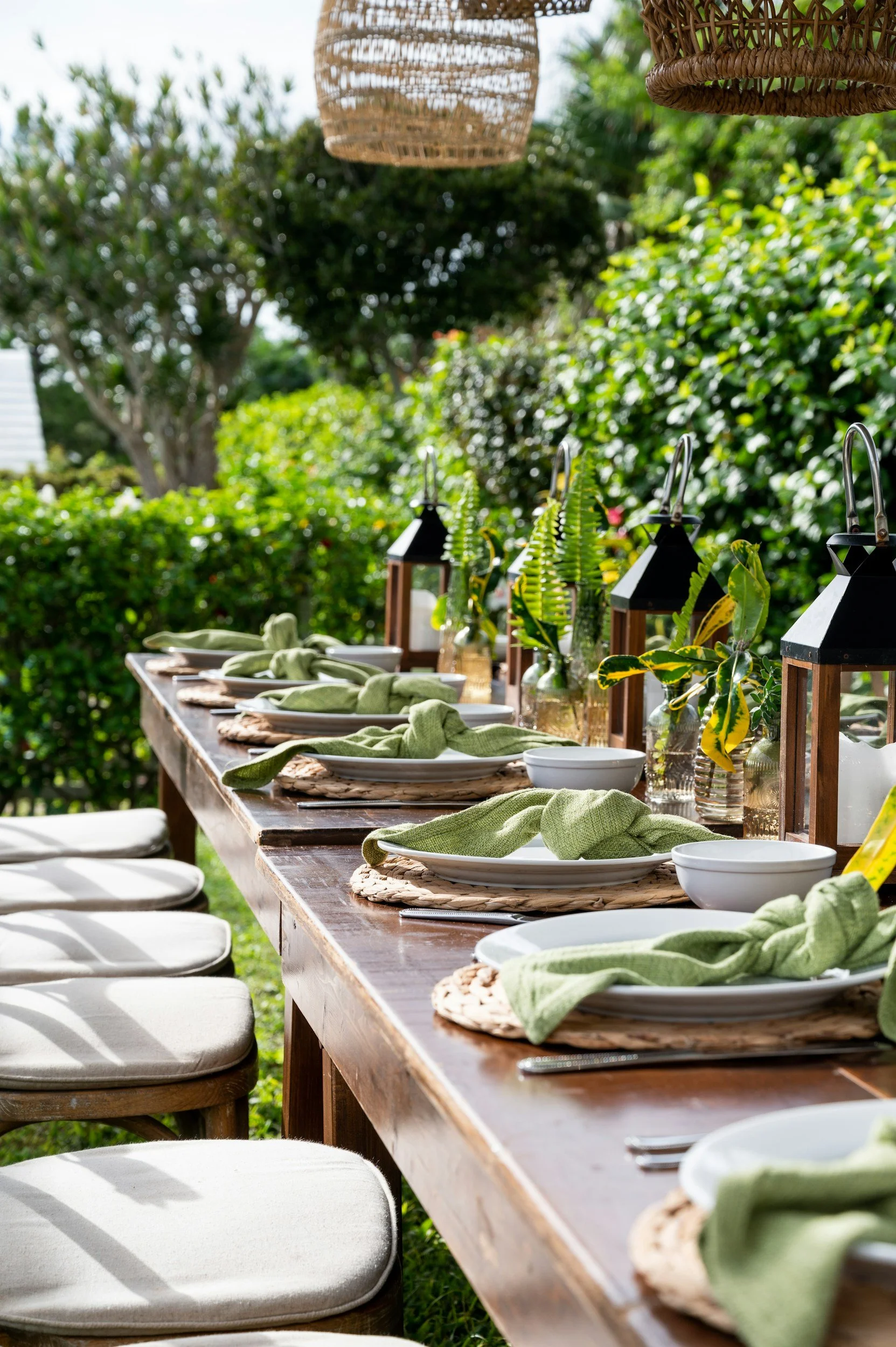 Outdoor dining table set for a gathering with white plates, green napkins, candles, lanterns, and lush greenery in the background.