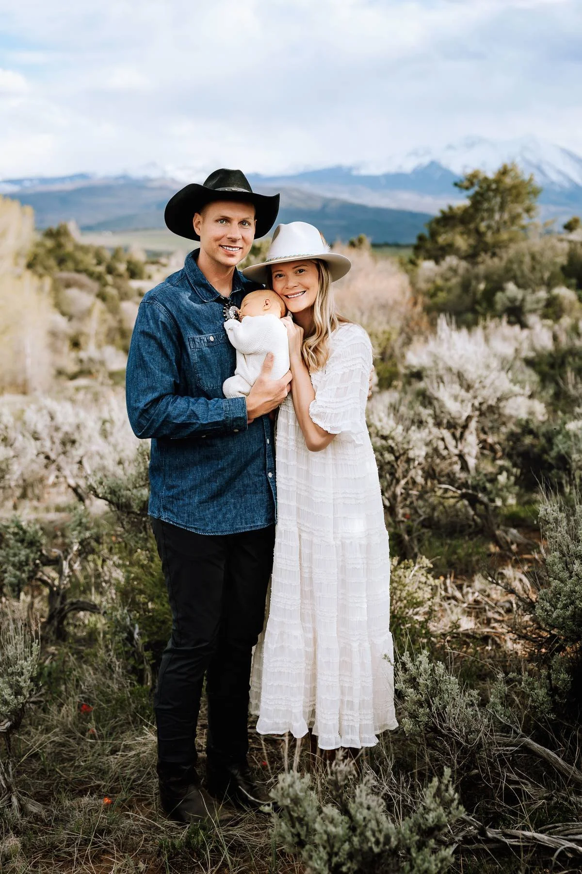 A family of three standing outdoors with mountains and cloudy sky in the background. The father wears a black cowboy hat and denim shirt, the mother wears a white hat and white dress, and the baby is wrapped in white clothing, sleeping in the mother's arms.