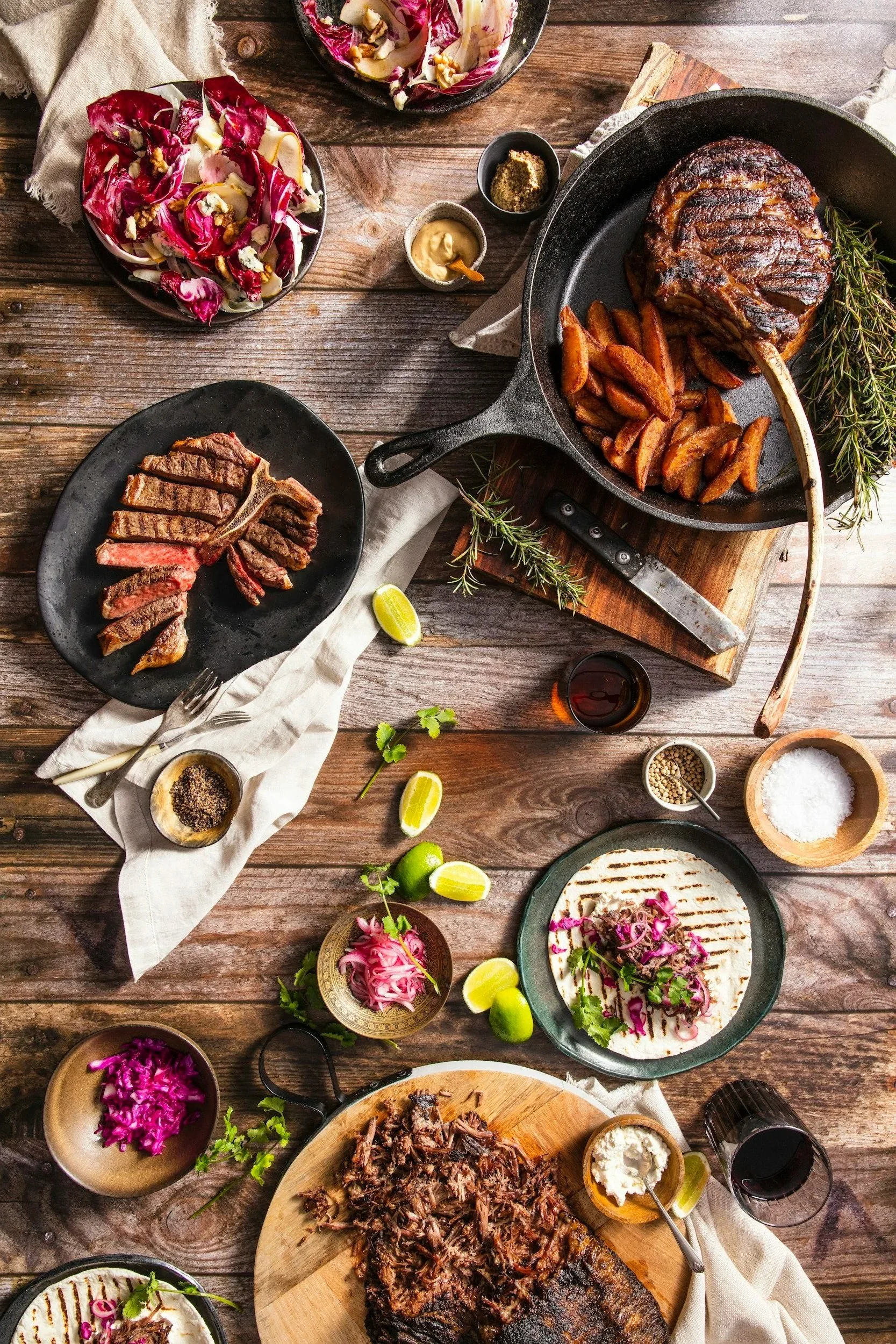 A rustic wooden table set for a meal featuring a large grilled steak, roasted potato wedges, and a cast iron skillet with a cooked meat or roast. The table includes bowls of colorful salads with purple and green vegetables, lemon wedges, various spices, and condiments, along with glasses of red wine and small bowls of pickled vegetables or garnishes.