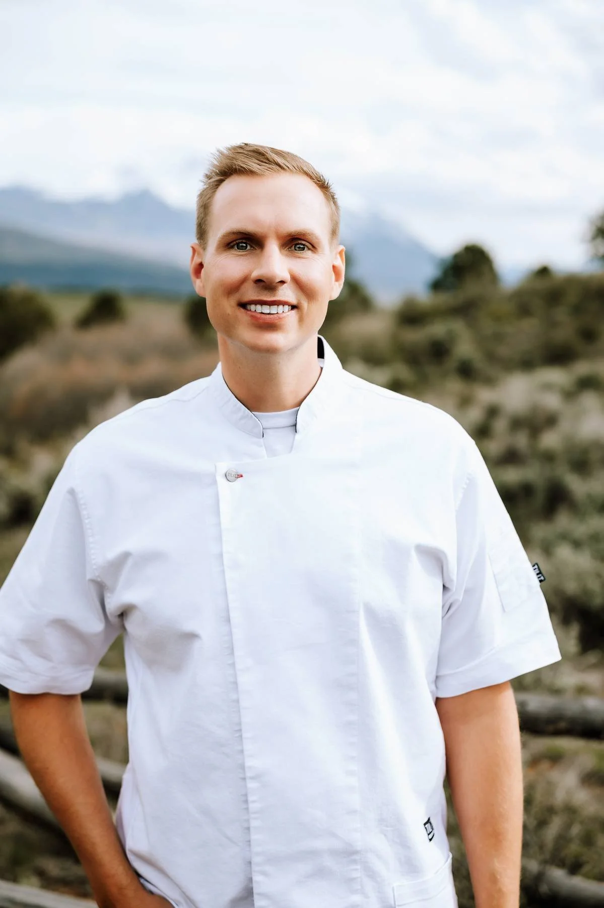 A young man in a white chef's coat standing outdoors with mountains and cloudy sky in the background.