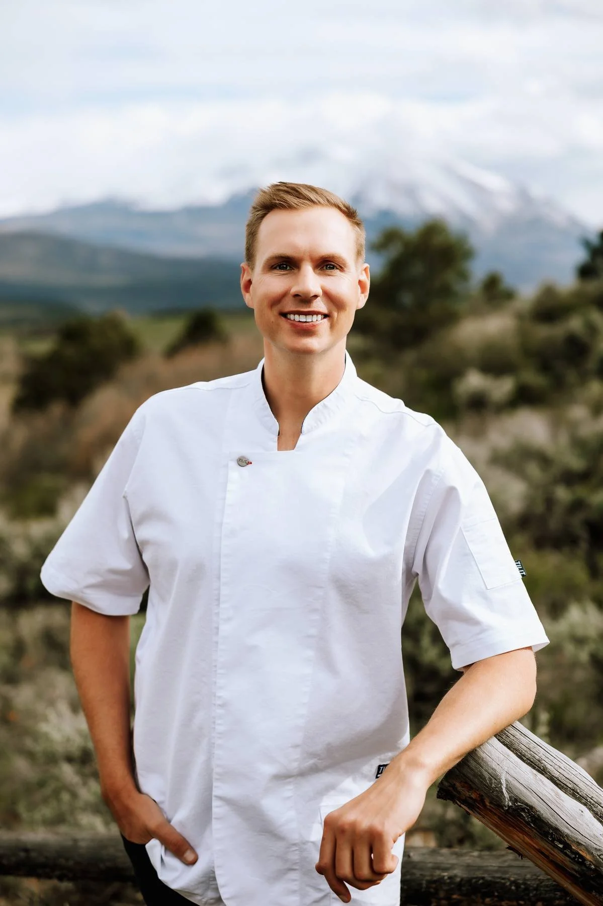 A smiling young man in a white chef's coat outdoors with a mountain and cloudy sky background.