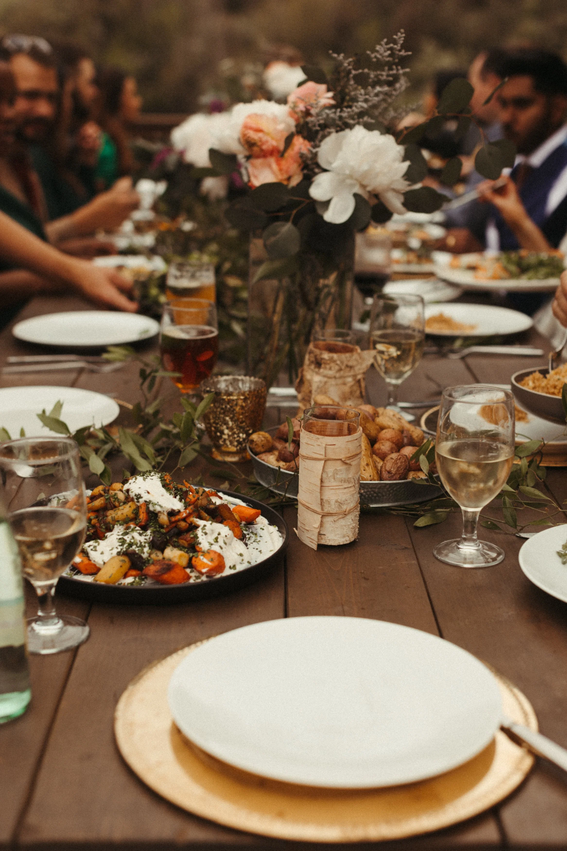 A table set for a meal with plates, glasses of wine, and a centerpiece of white and peach flowers with greenery. The table has various dishes including roasted vegetables and bread rolls, and people are gathered around.