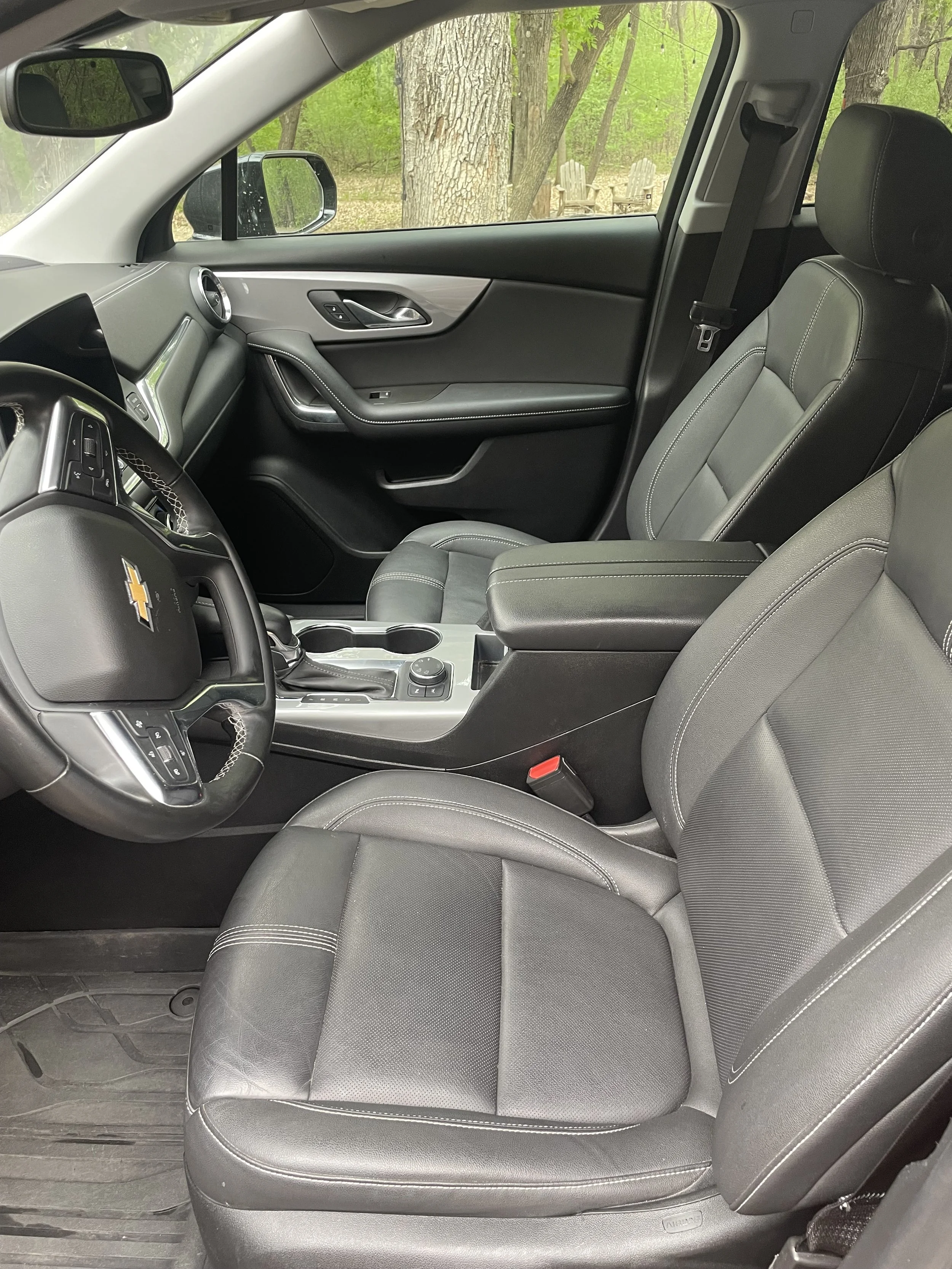 The interior of a Chevrolet vehicle showing the front passenger seat, dashboard, and steering wheel, with a view of trees and outdoor chairs through the window.
