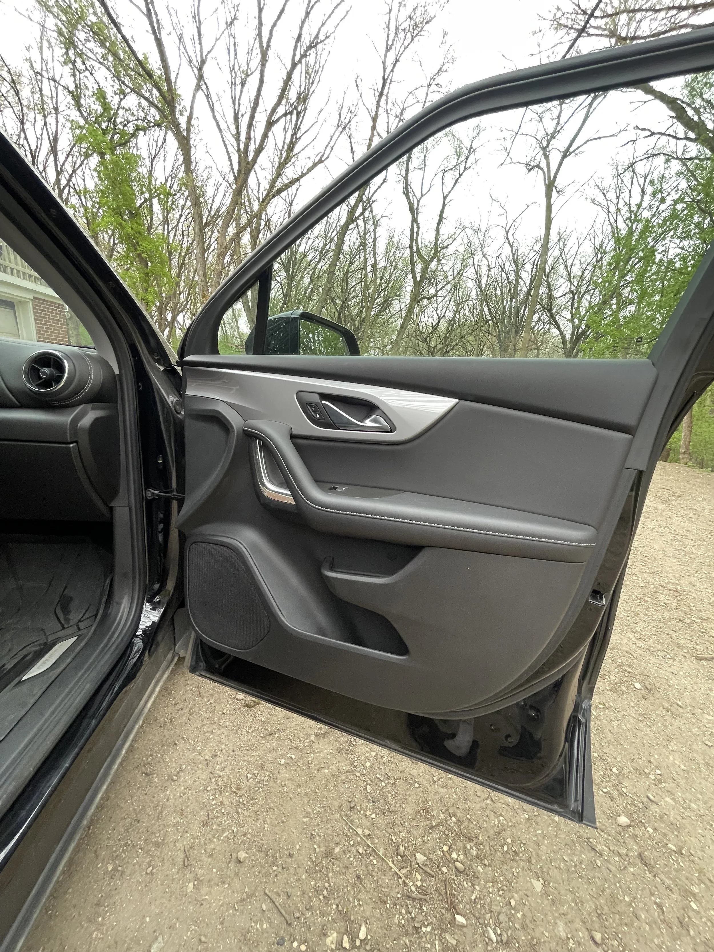 Open driver side door of a black car, showing the interior door panel, parked on dirt and gravel with leafless trees in the background.