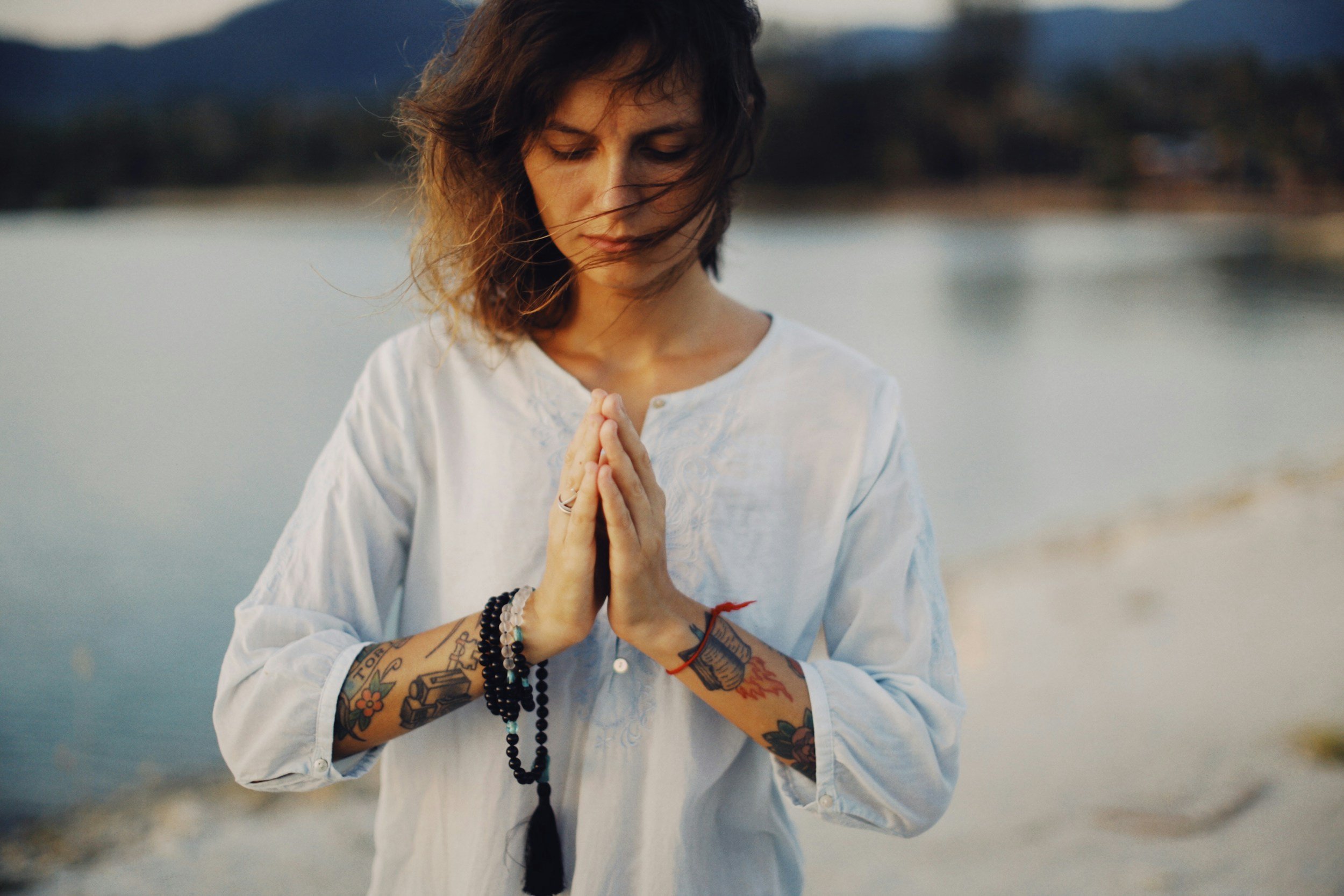 A woman with dark, wavy hair and tattoos on her arms is standing outdoors near a body of water, with her eyes closed and hands pressed together in prayer or meditation.
