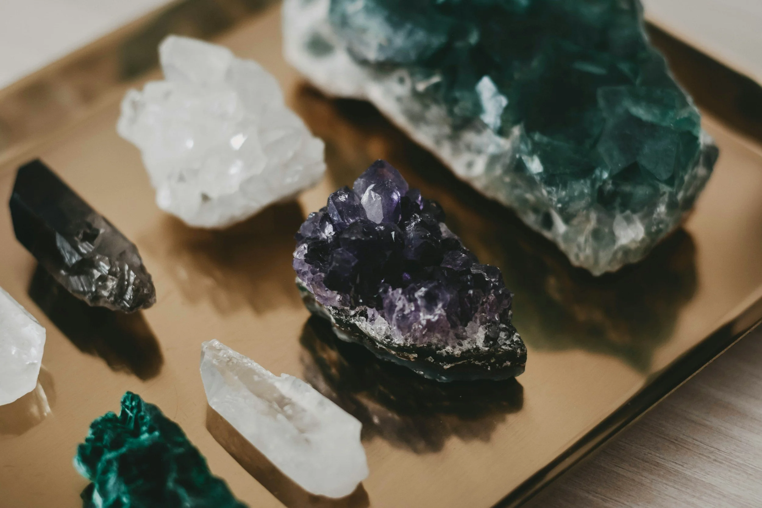 Various mineral and crystal specimens displayed on a wooden surface, including an amethyst cluster, green stone, white mineral, and dark metallic mineral, with some rough and polished appearances.