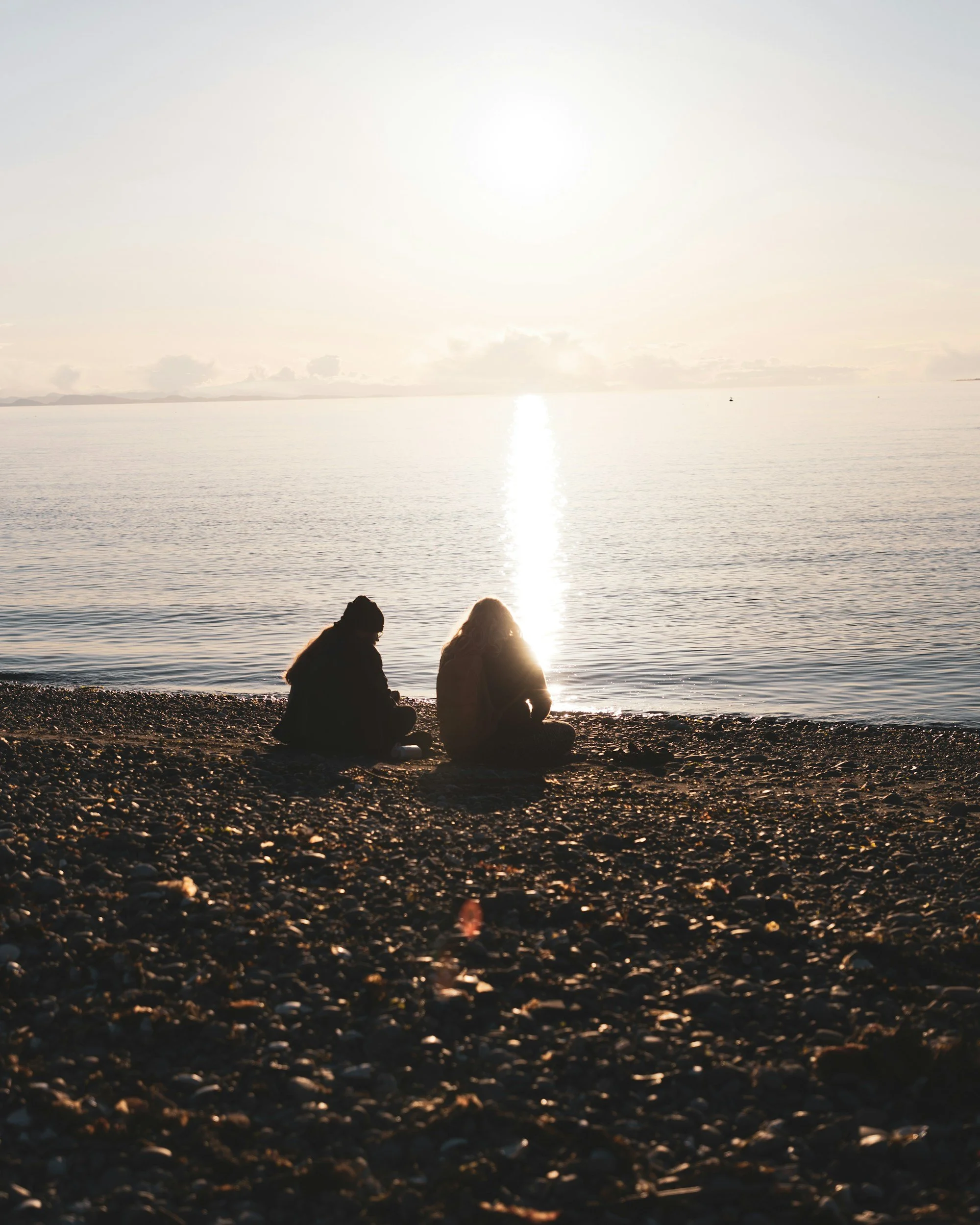 Two people sitting on a rocky beach during sunset or sunrise, facing the water with the bright sun reflecting on the calm sea.