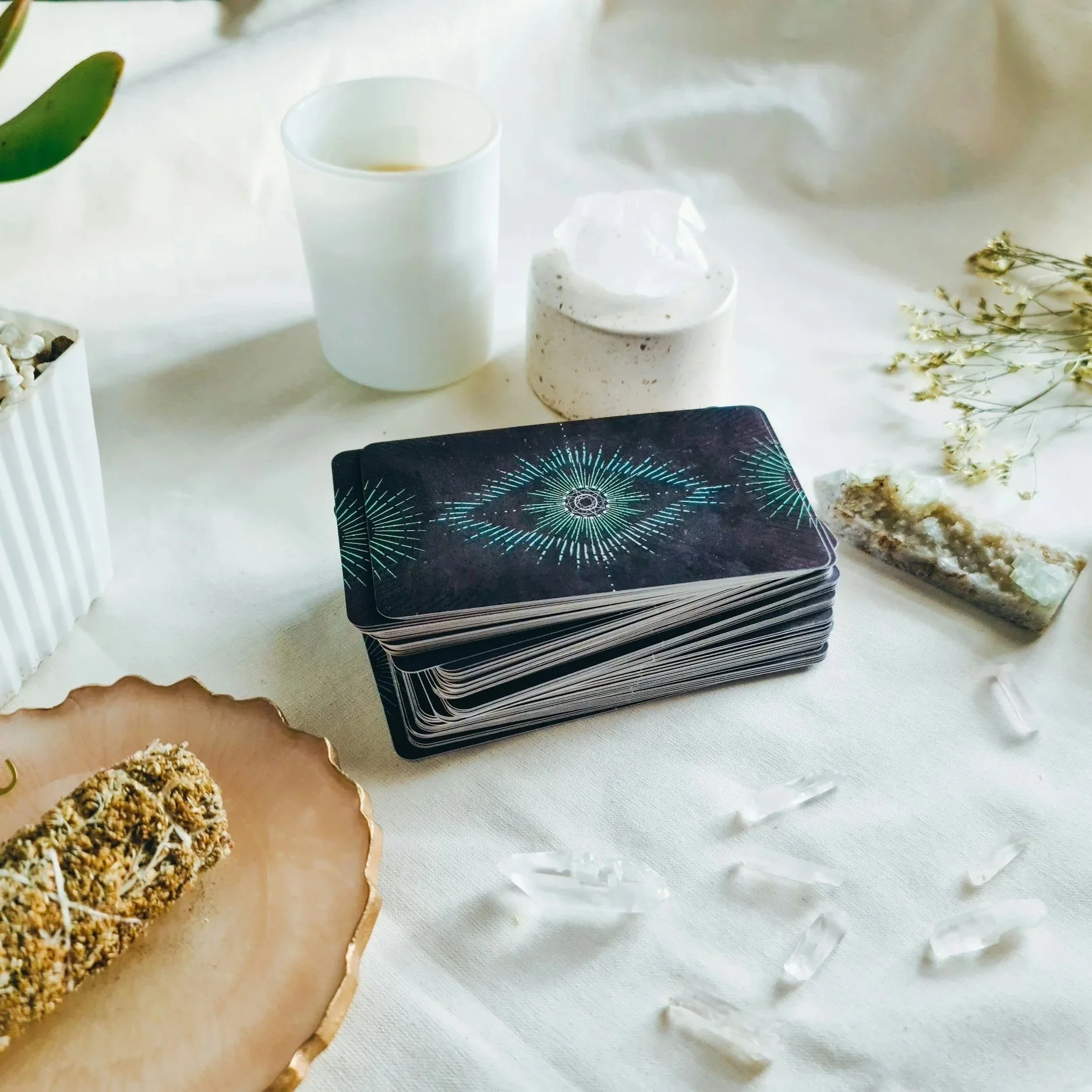 A tarot card deck with geometric pattern on a white table, alongside a cup of tea or coffee in a white mug, a small white speckled ceramic container with a clear lid, a golden-rimmed plate with a baked good topped with a crumble, and a sprig of small white flowers.