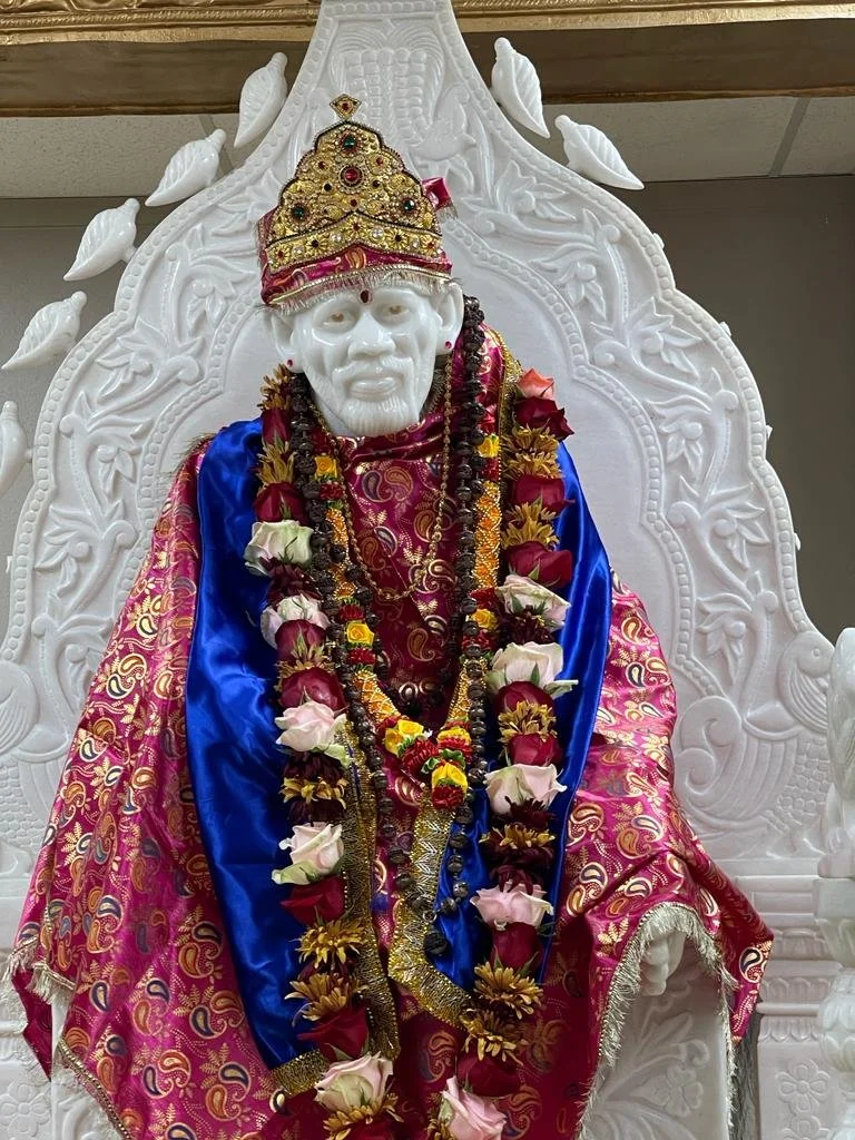 A white statue of Shirdi Sai Baba, wearing a decorated crown, and adorned with multiple flower garlands, sitting on an ornate white throne with detailed carvings.