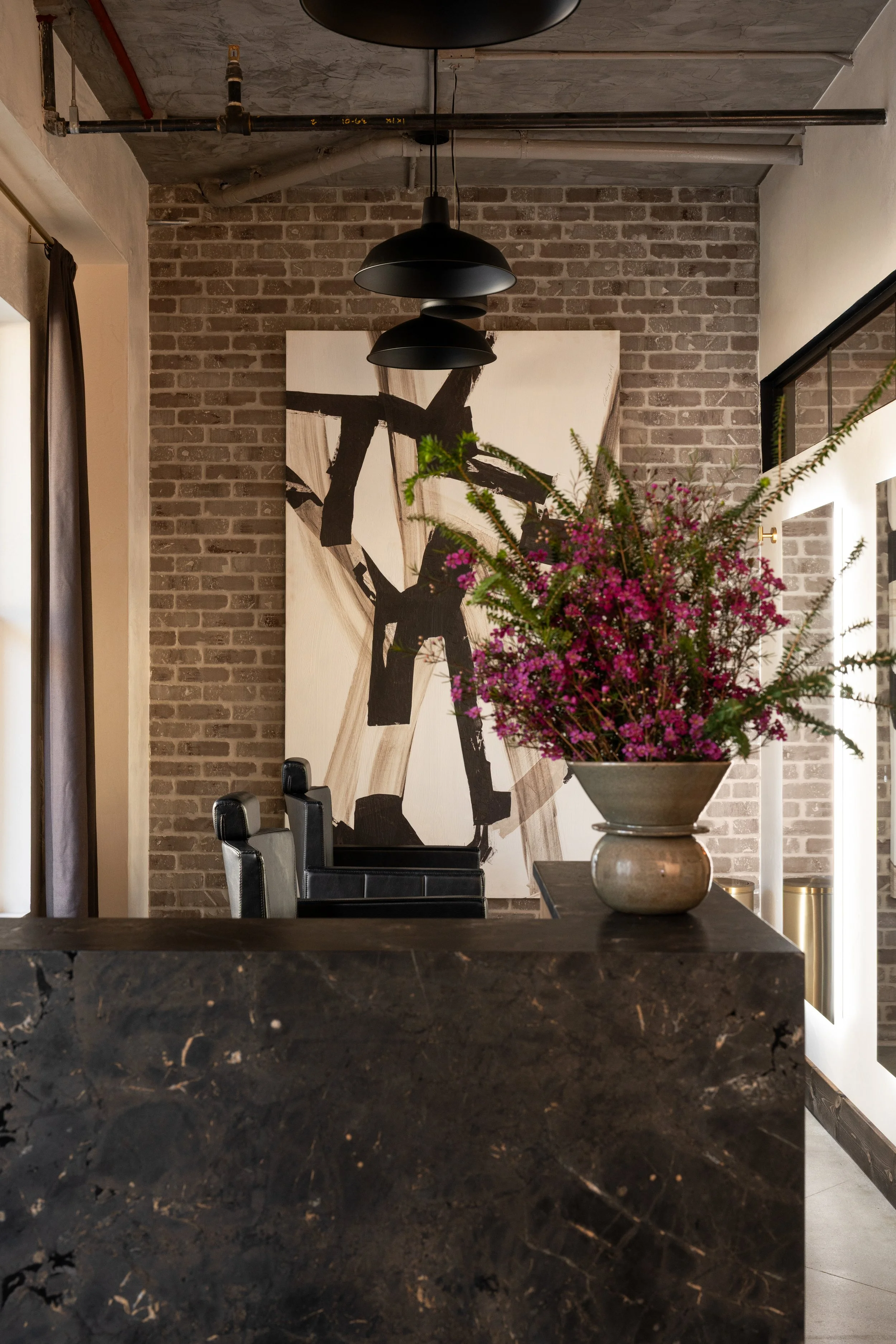 Interior of a modern dining or conference room with exposed brick wall, large black and white abstract artwork, a black leather chair, and a black table with a floral arrangement of pink and green flowers in a metallic vase.