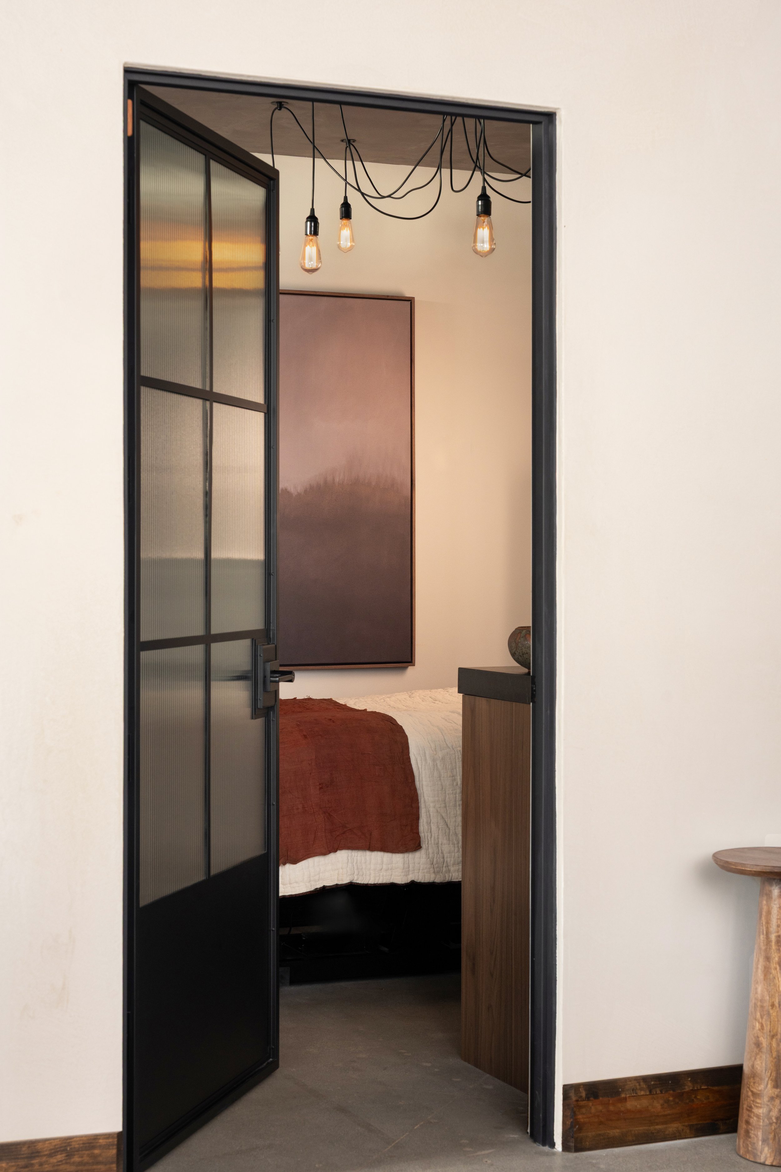 A black-framed glass door opened into a bedroom with a bed, rust-colored blanket, beige wall, and hanging Edison-style light bulbs.