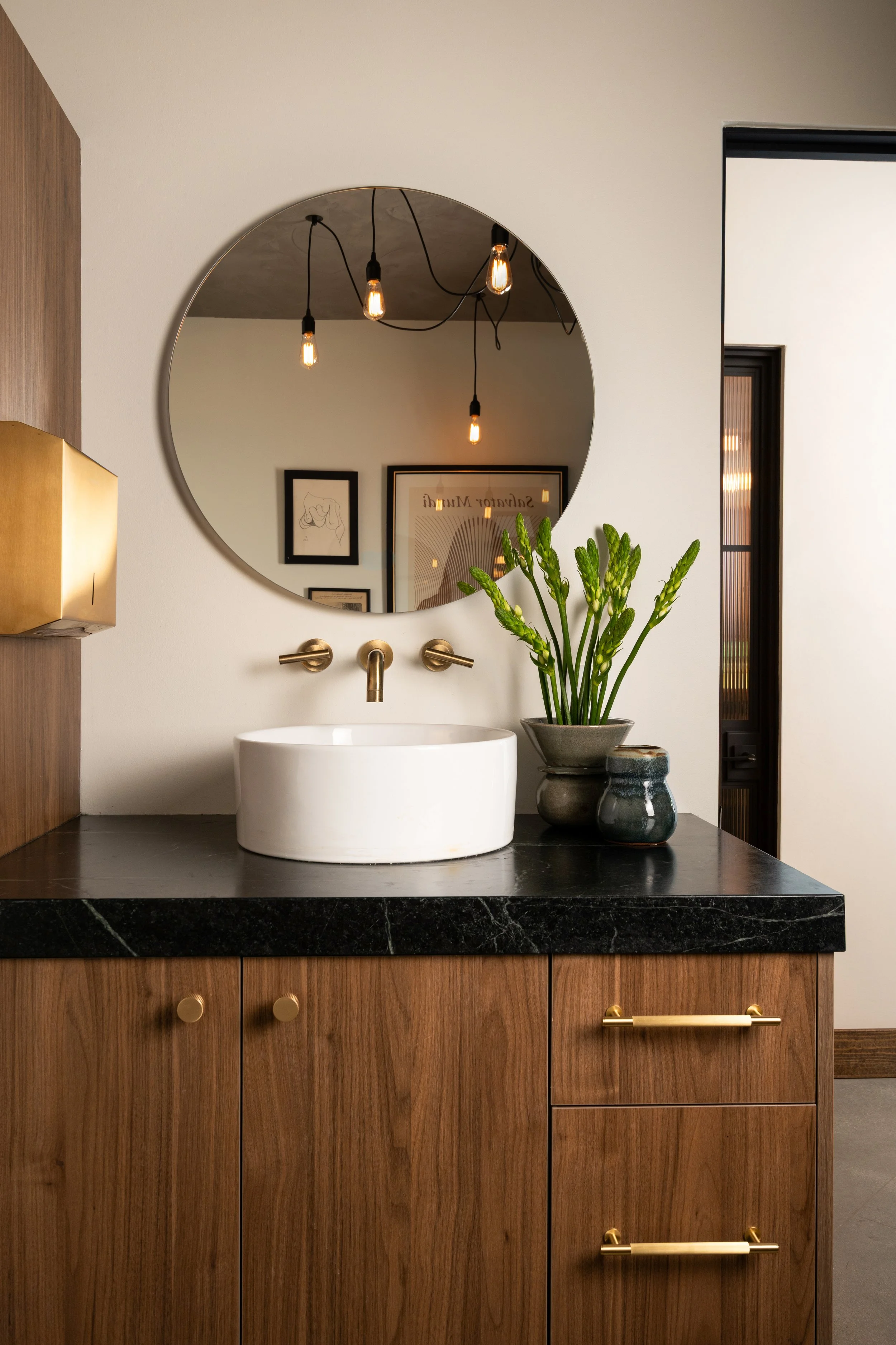Bathroom vanity with a round mirror, a white vessel sink, gold fixtures, potted green plant, framed artwork on the wall, and wooden cabinets with gold handles.