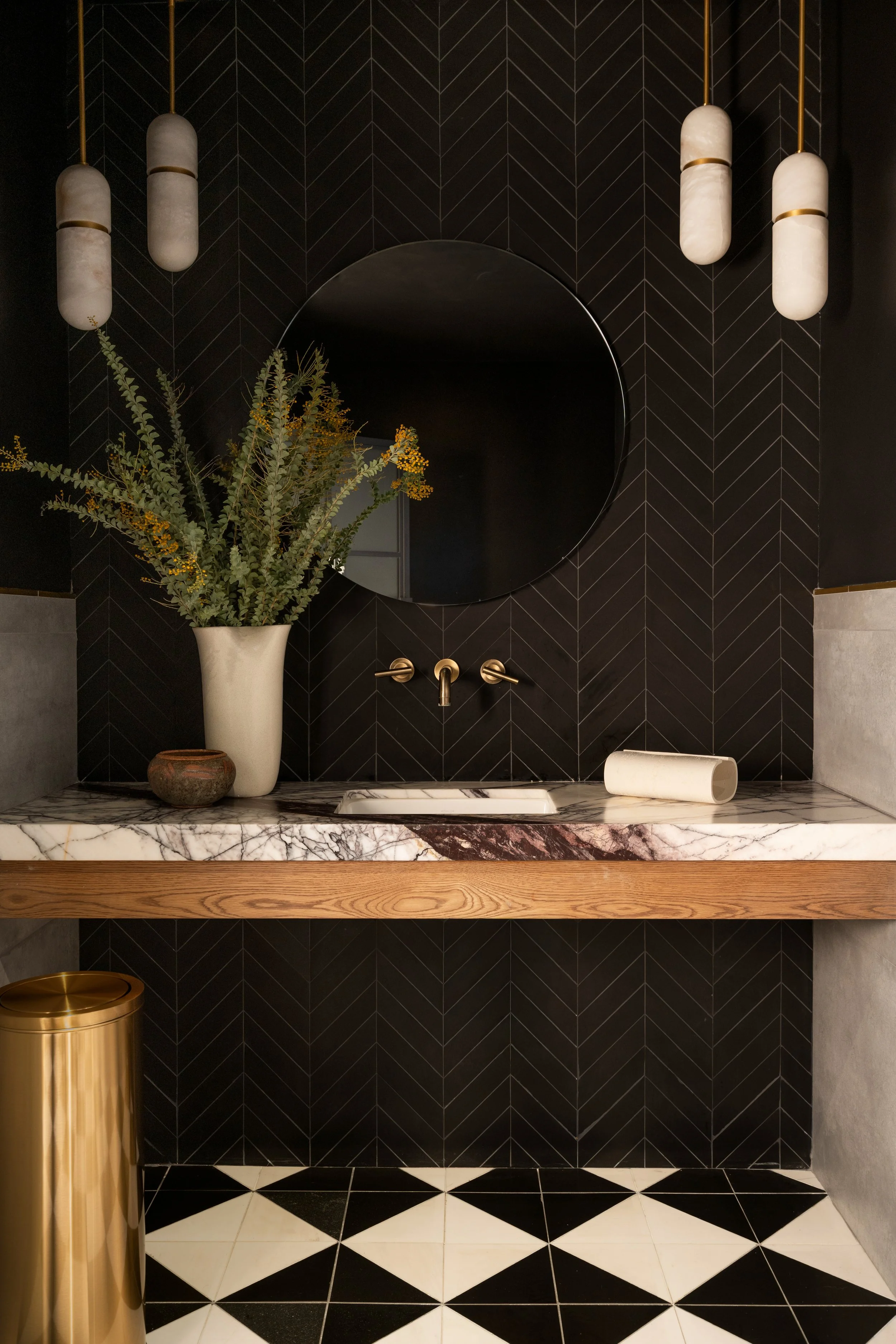 Modern bathroom with black herringbone wall tiles, a round mirror, a marble countertop with a sink, a tall white vase with greenery, a small decorative bowl, and cylindrical wall lights with brass accents.
