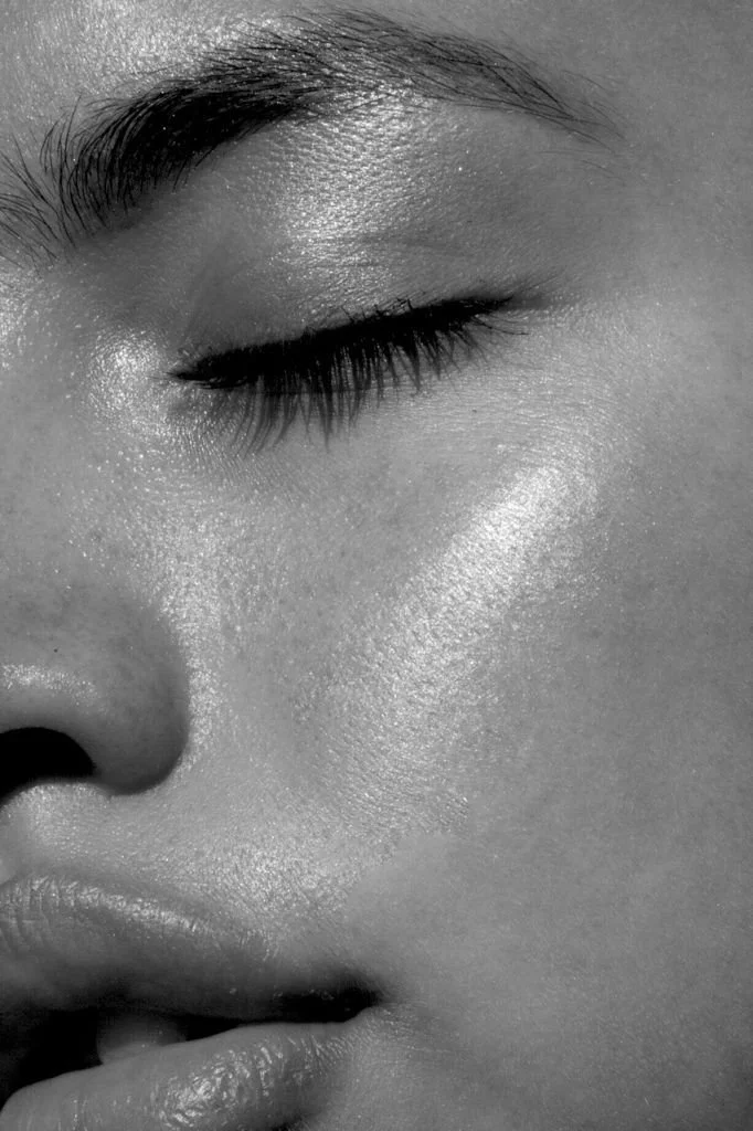 Close-up black and white photo of a person's face with their eyes closed, showing detailed skin texture, eyelashes, and eyebrow.