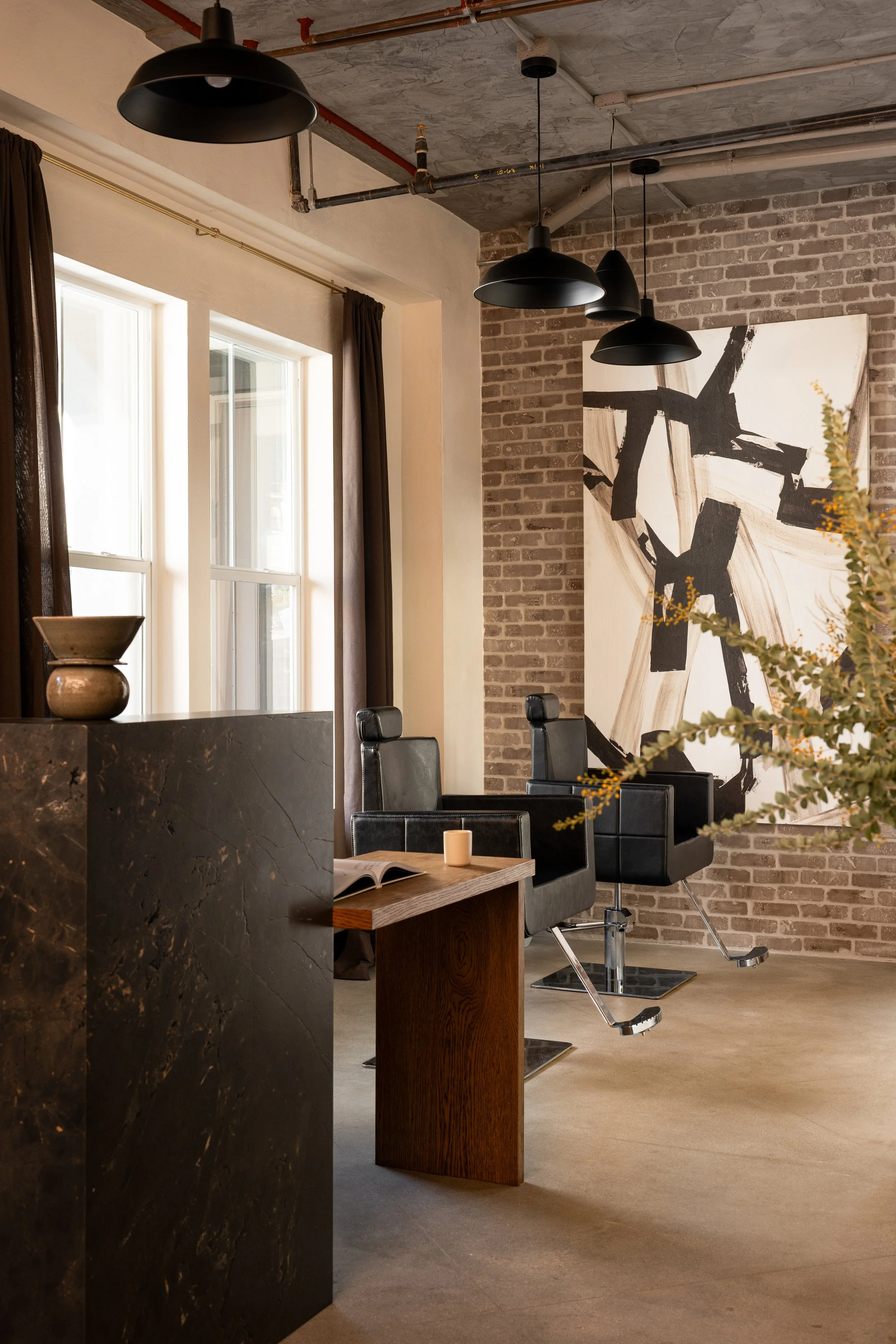A modern salon interior with black salon chairs, a computer, a wooden side table, a large abstract black and white painting on a brick wall, and hanging black pendant lights. Sunlight streams in through tall windows covered with dark curtains.