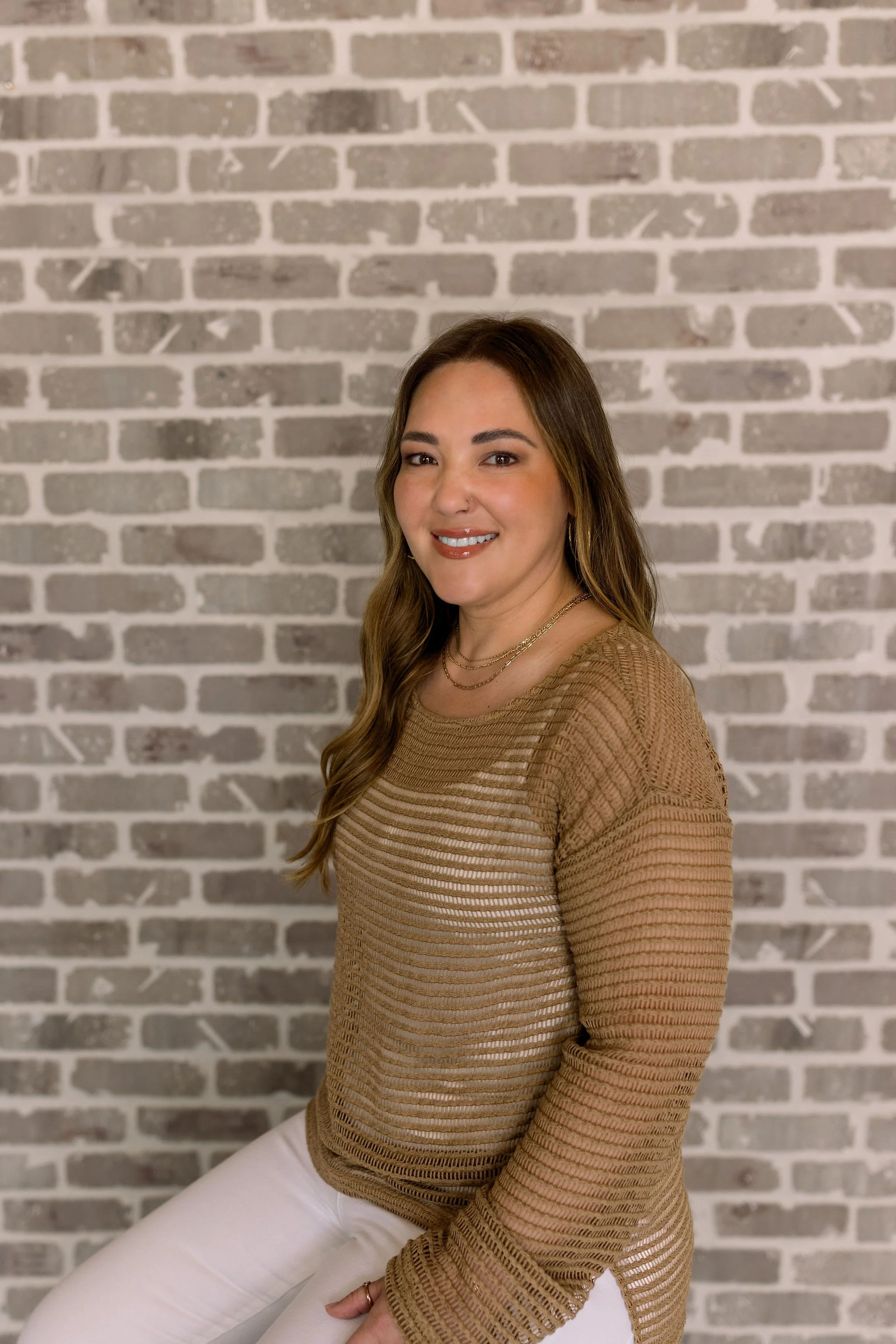 A woman with long wavy brown hair, wearing a semi-transparent tan striped long-sleeve top and white pants, sitting against a gray brick wall, smiling and looking at the camera.