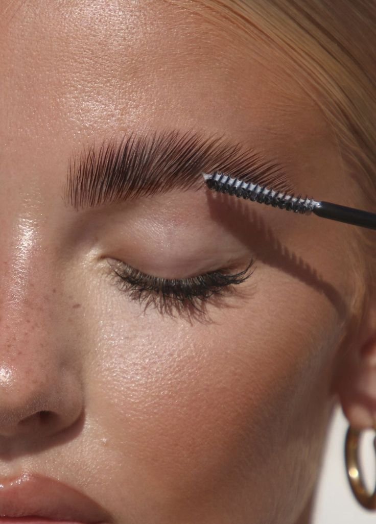 Close-up of a person with light skin, applying mascara to their upper eyelashes using a mascara wand, with well-groomed eyebrows.