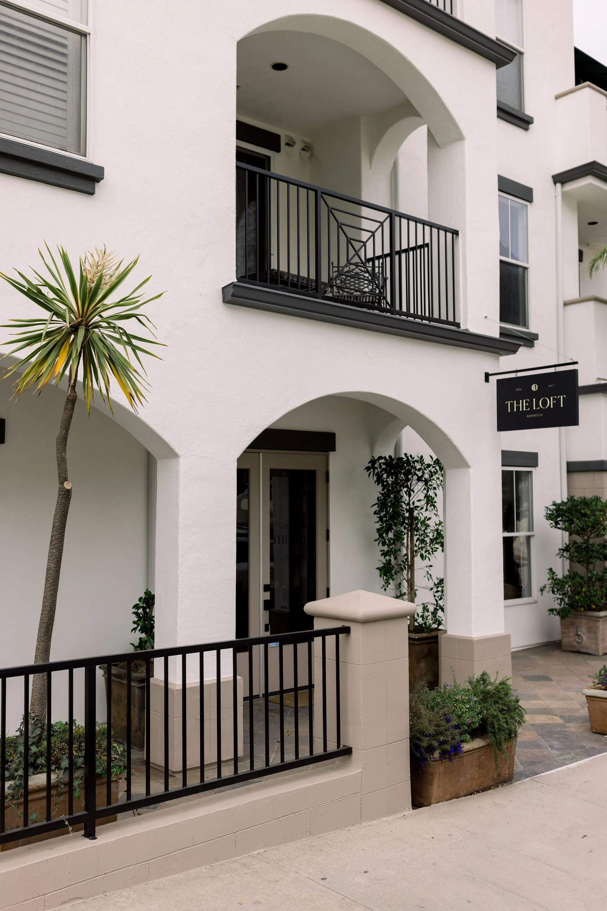 Exterior view of a modern white multi-story building with black accents, featuring a small balcony with black metal railing, a sign that reads 'The Loft,' and various potted plants and small trees in front.