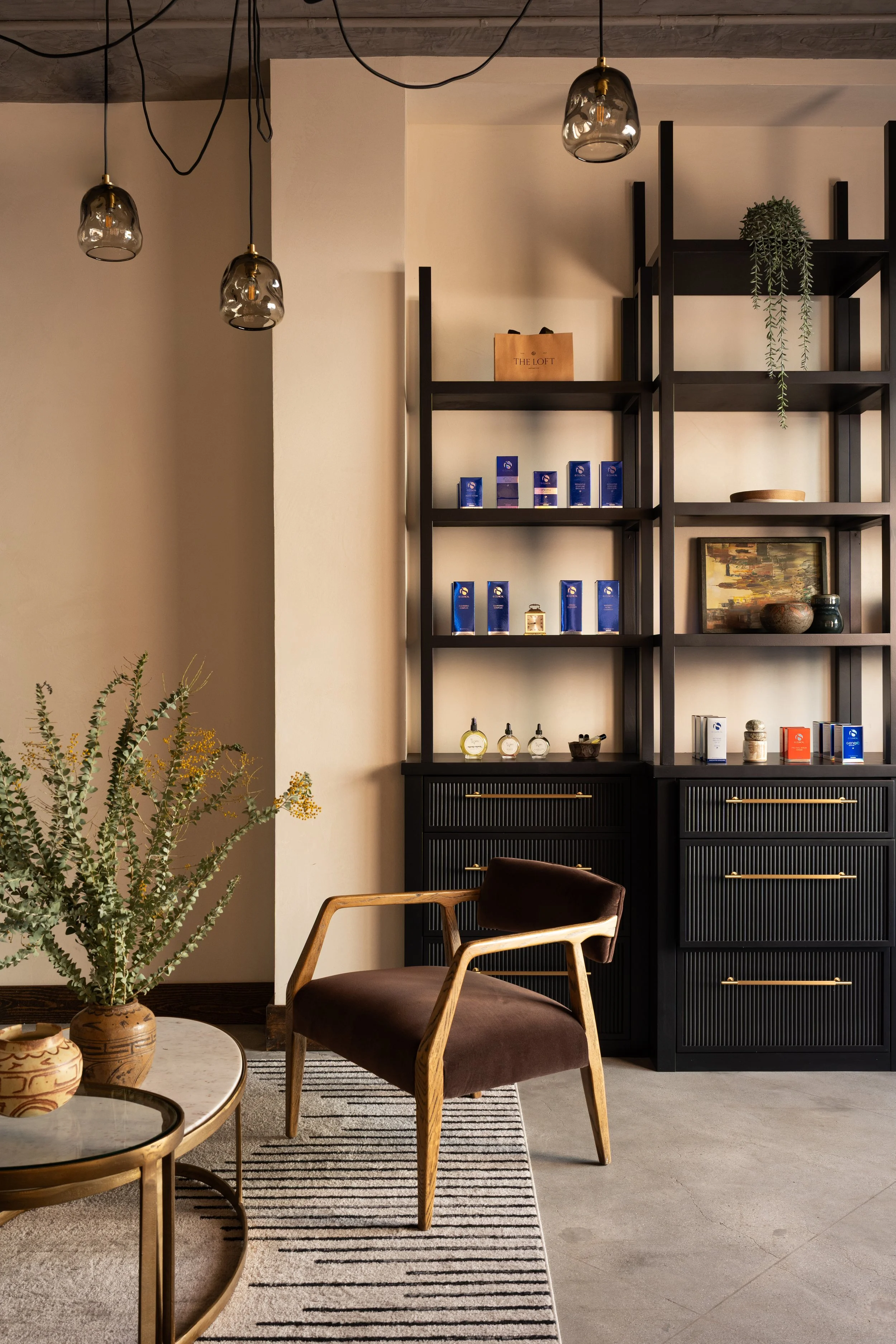 Modern interior with black shelving unit, decorative items, and plants. Brown chair in foreground, round coffee table with pottery, and striped rug on concrete floor. Pendant lights hanging from ceiling.