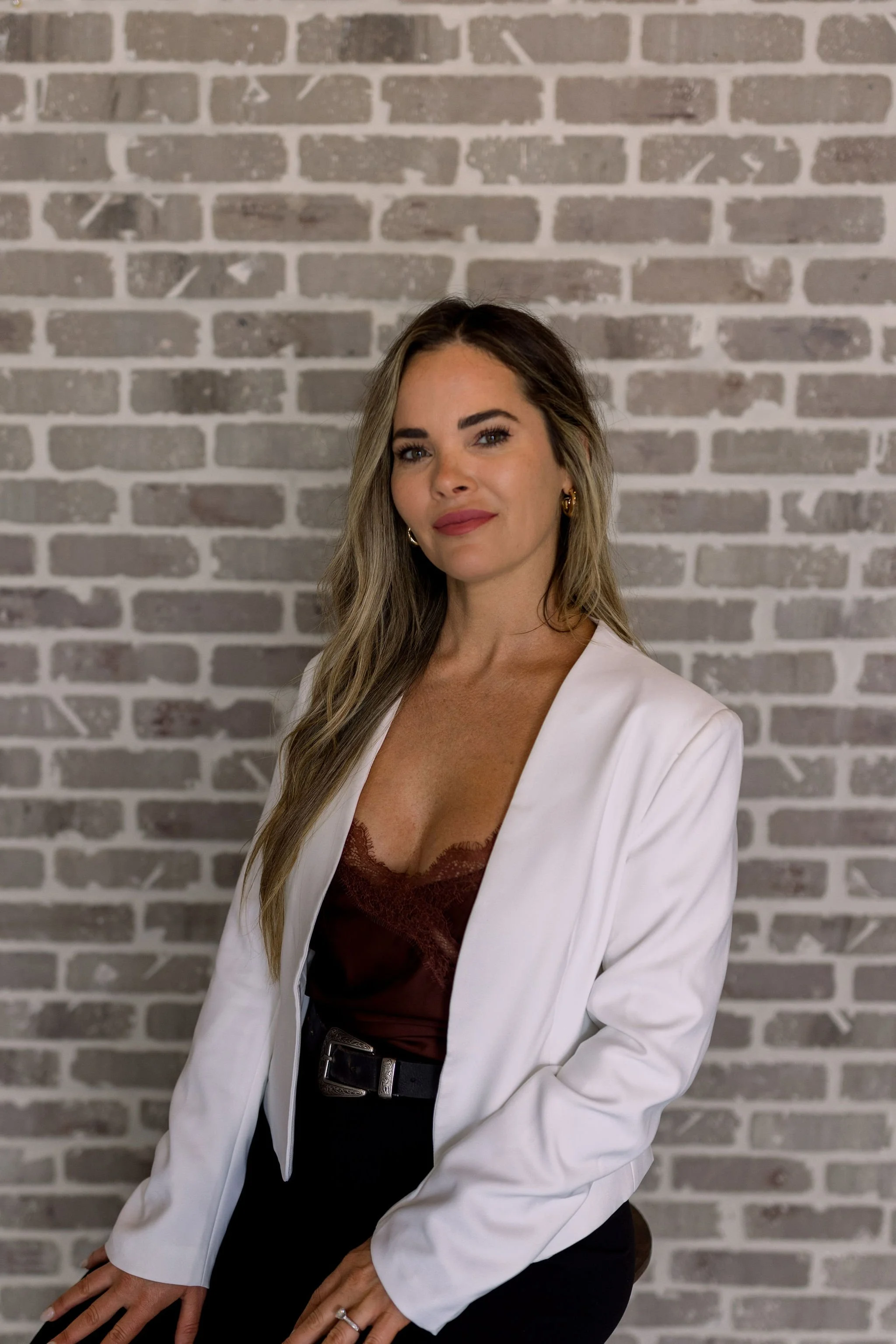 A woman with long wavy light brown hair and fair skin is sitting in front of a gray brick wall. She is wearing a white blazer, a maroon lace top, and black pants with a belt. She has earrings and a ring, and is looking slightly to her right with a slight smile.