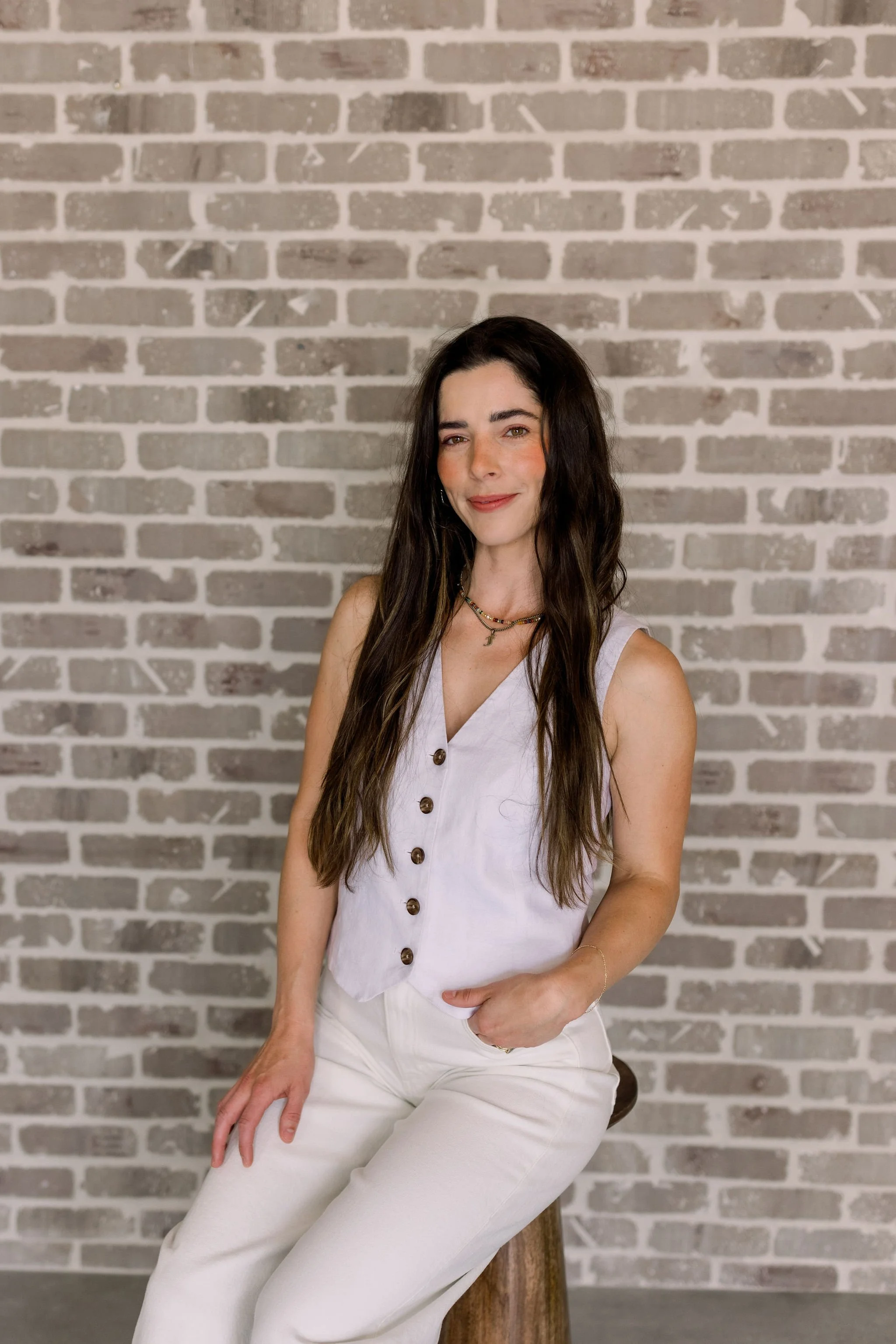 A woman with long dark hair and a light skin tone, sitting on a wooden stool in front of a gray brick wall. She is wearing a sleeveless white button-up top and white pants, accessorized with layered necklaces and a bracelet. She has a slight smile on her face.