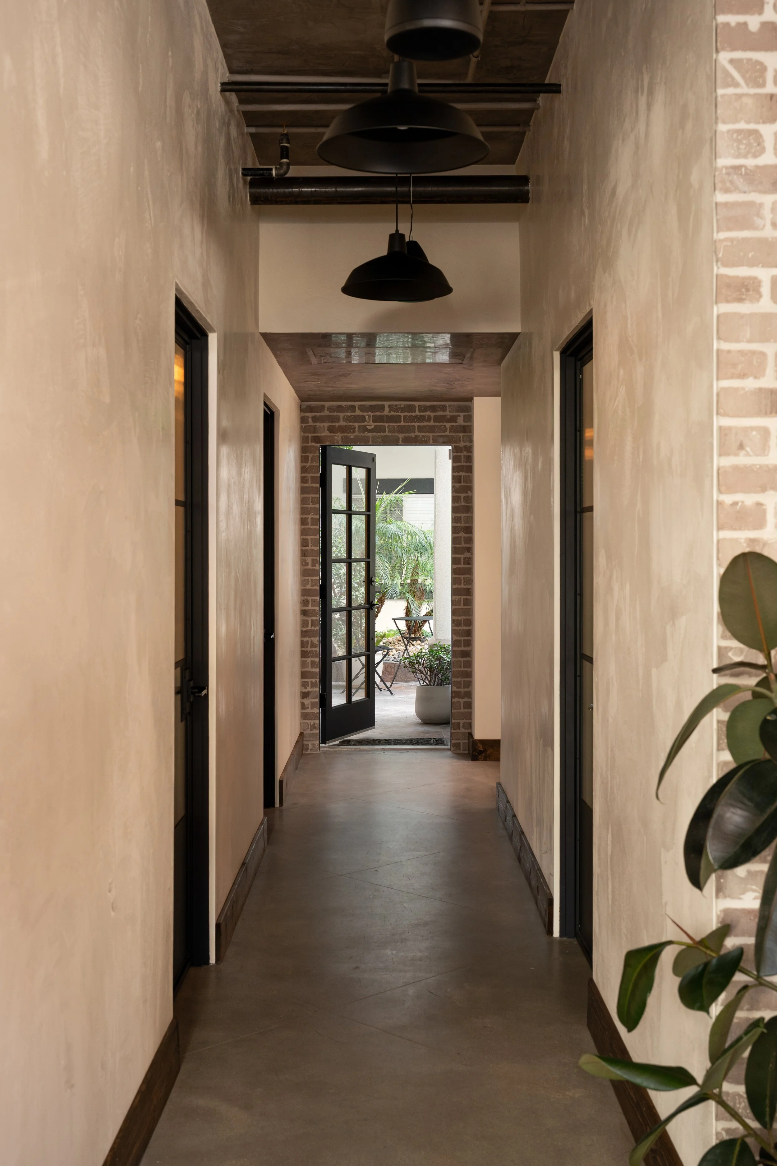 A corridor with a concrete floor, beige walls, and black-framed doors on each side. At the end is a doorway with a glass door opening to an outdoor patio with plants, and two black pendant lights hang from the ceiling.