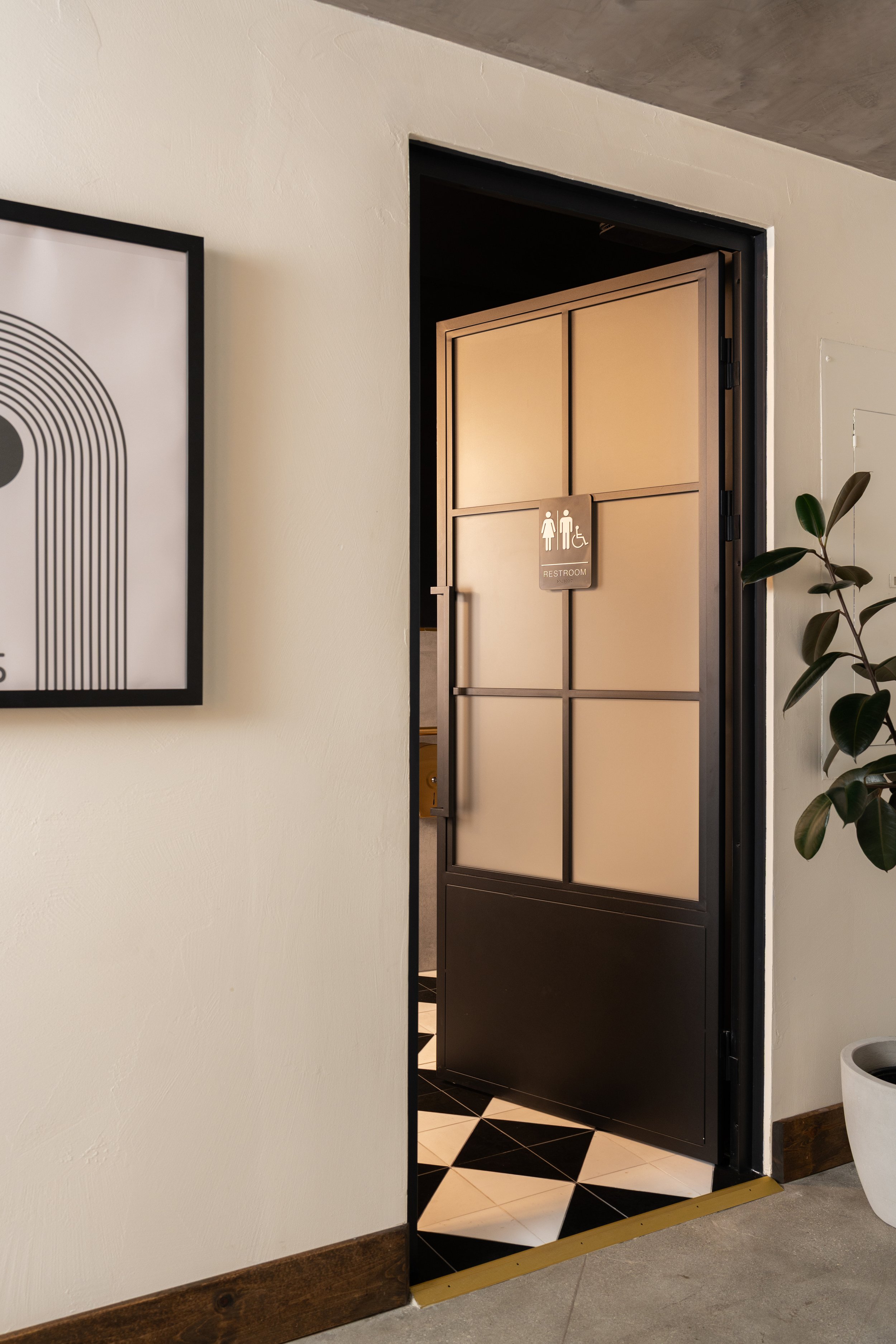 Bathroom door with a sign indicating accessible restroom, next to a plant and art on the wall, with black and white checkered floor tiles.