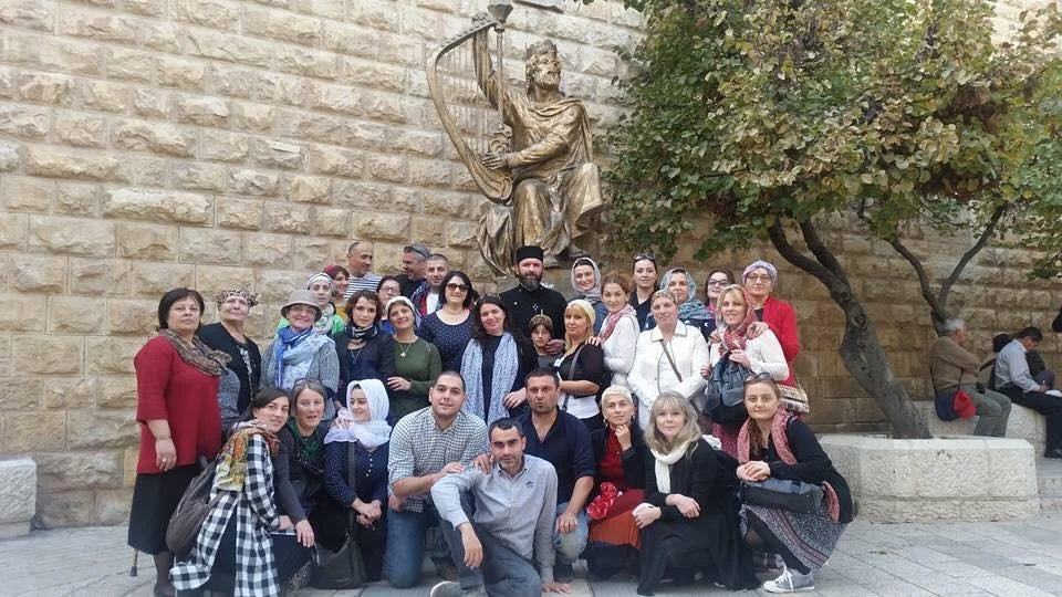 Grupo de personas posando en frente de una estatua y un muro de piedra, algunos sentados y otros de pie.