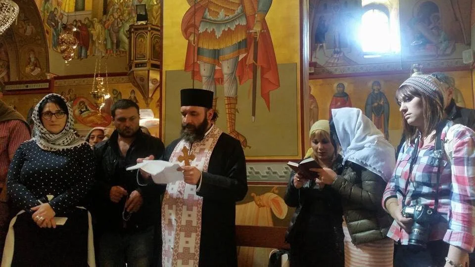 Grupo de personas en una iglesia ortodoxa, participando en una ceremonia religiosa, con un sacerdote en el centro leyendo un libro, rodeado de fieles y iconos religiosos en las paredes.