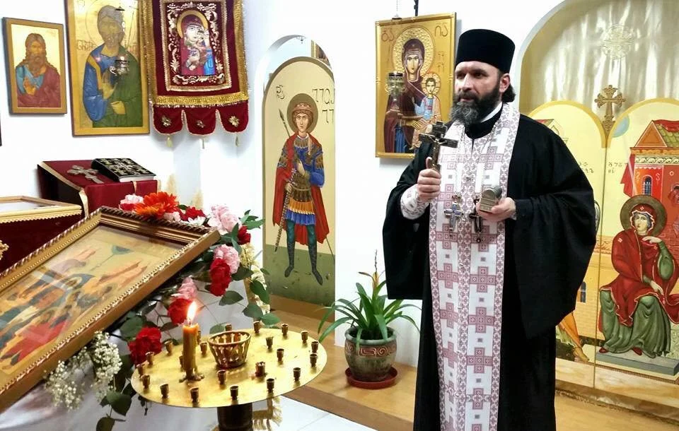 Un sacerdote ortodoxo en una iglesia, sosteniendo una cruz y un libro, rodeado de iconos religiosos y flores en un altar.