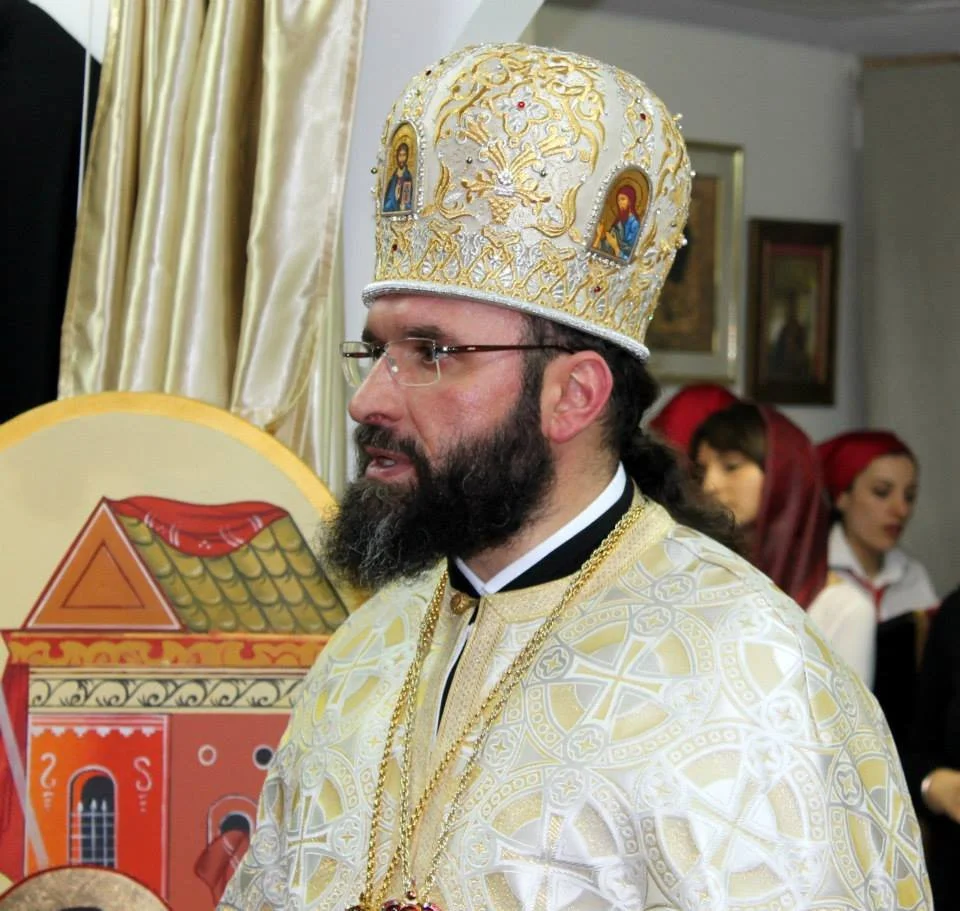 Hombre con barba, gafas y vestimenta religiosa, lleva mitra adornada con íconos religiosos y detalles dorados, en un entorno con otras personas vestidas con ropa tradicional.