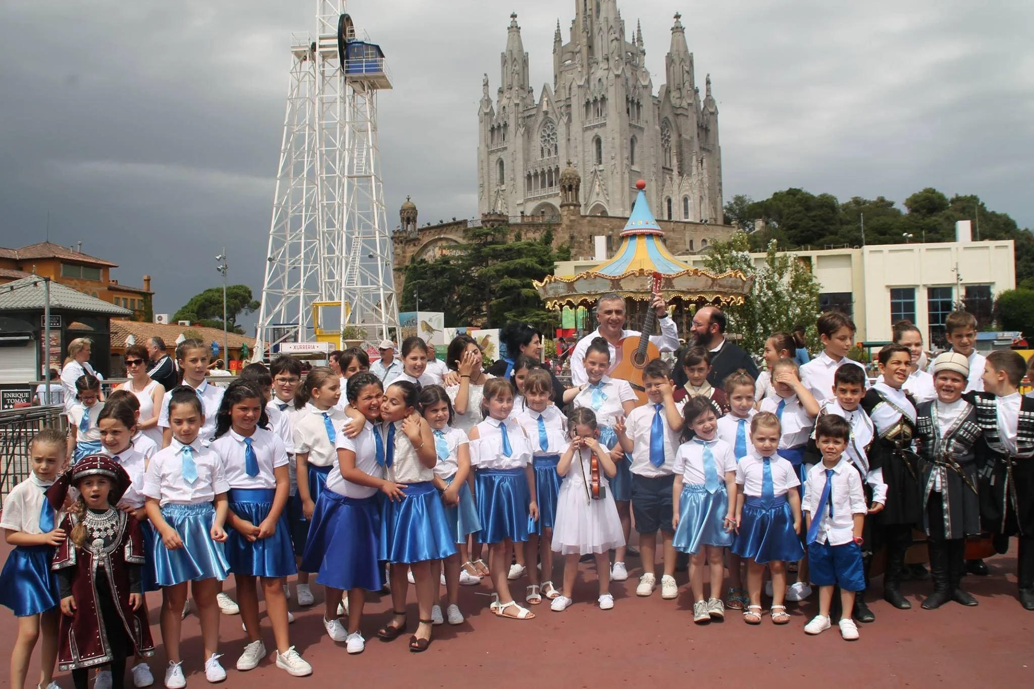 Un grupo de niños y adultos en una plaza, algunos con ropa típica, rodeando a una persona que toca guitarra, con el Castillo de Chapultepec y una noria en el fondo, en un día nublado.