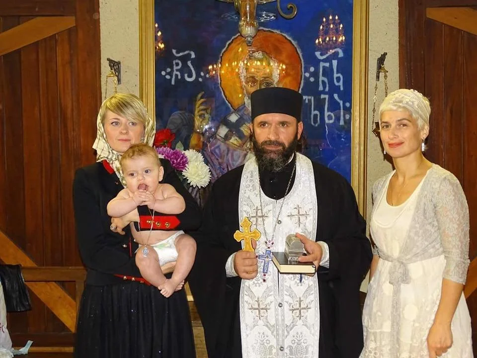 cuatro personas en una ceremonia religiosa, un sacerdote con un niño en brazos, una mujer con cabello rubio y vestido blanco, y otra mujer con cabello rubio y vestido negro, en un fondo con icono religioso
