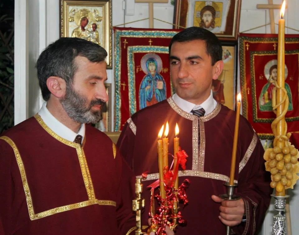 Dos hombres en vestimenta litúrgica sosteniendo velas y en un entorno de iconos religiosos