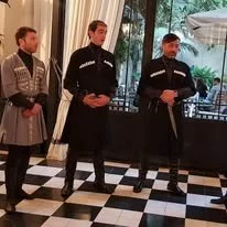 Tres guardias de seguridad en un piso de baldosas negras y blancas, con fondo de ventanas con vista a árboles y un toldo blanco.