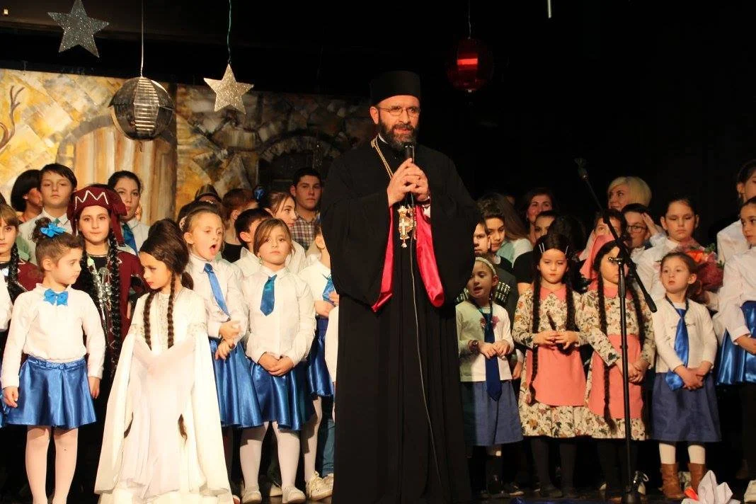 Un sacerdote con barba y lentes usando vestimenta clerical, con un micrófono en mano, frente a un grupo de niños en uniforme y vestimenta tradicional, en un escenario decorado con estrellas y esferas plateadas, durante un evento o presentación.