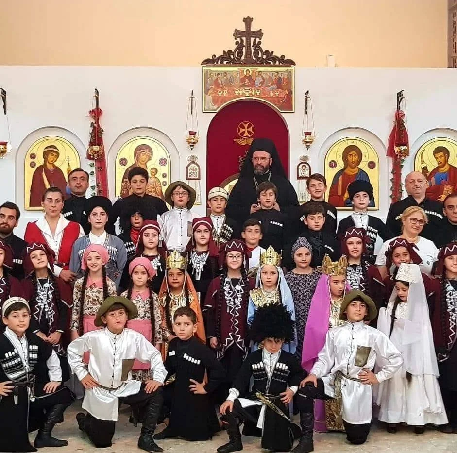 Grupo de niños y adultos en un templo ortodoxo, vestidos con trajes tradicionales y religiosos, posando para una foto en el altar con iconos religiosos de fondo.