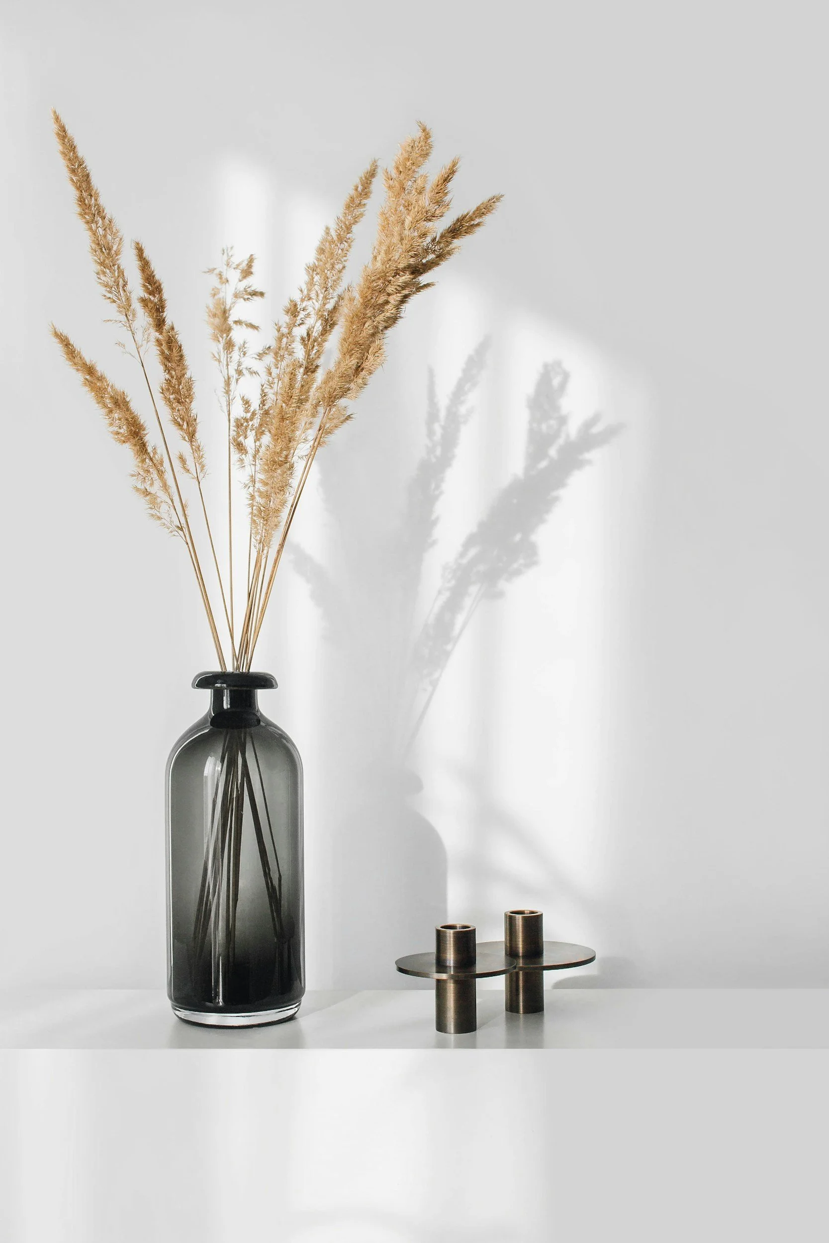 A minimalist still life with a black glass vase holding dried beige pampas grass. Shadows cast on a white wall behind. To the right, a small brass round table holding three matching brass candle holders.