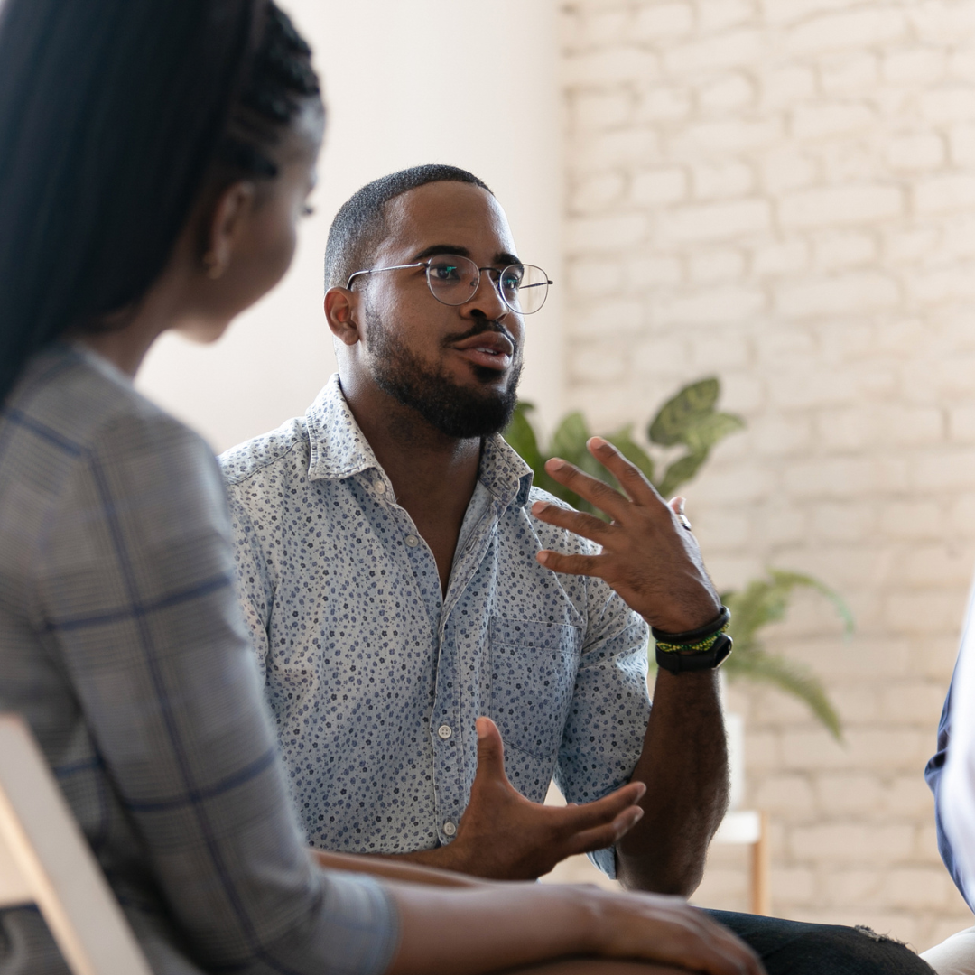 Two people having a conversation in an indoor setting with a brick wall background and a green plant.