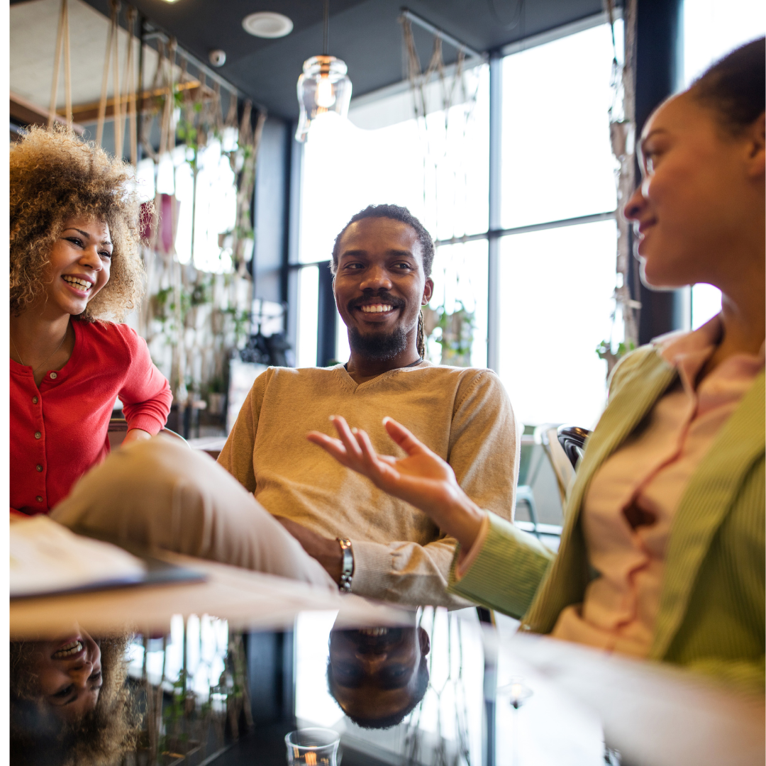 Three friends, two women and a man, having a conversation and smiling in a brightly lit cafe with large windows and hanging plants.