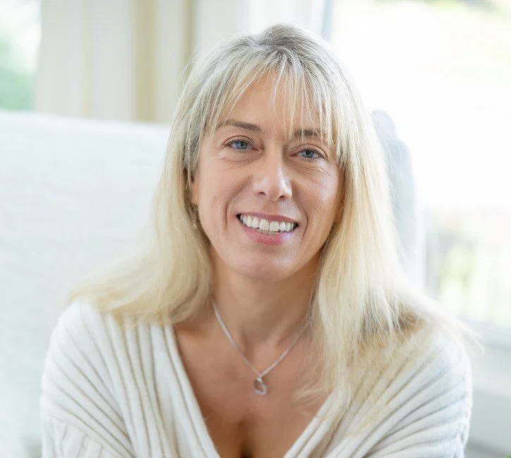 A blonde woman with a big smile, wearing a white striped top and a silver necklace, sitting indoors near a window with greenery outside.