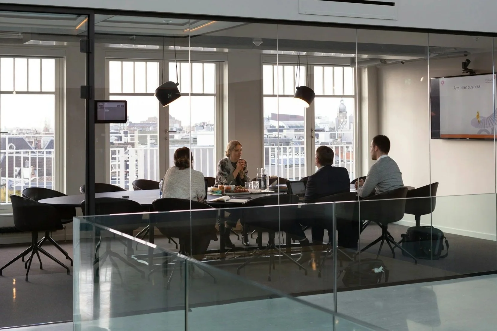 Four people sitting around a conference table in a modern office with large windows, two women and two men, with a cityscape view outside.