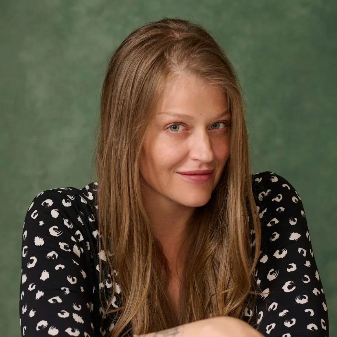 A person with long, wavy, light brown hair and green eyes smiling at the camera, wearing a black shirt with a white pattern, against a green background.