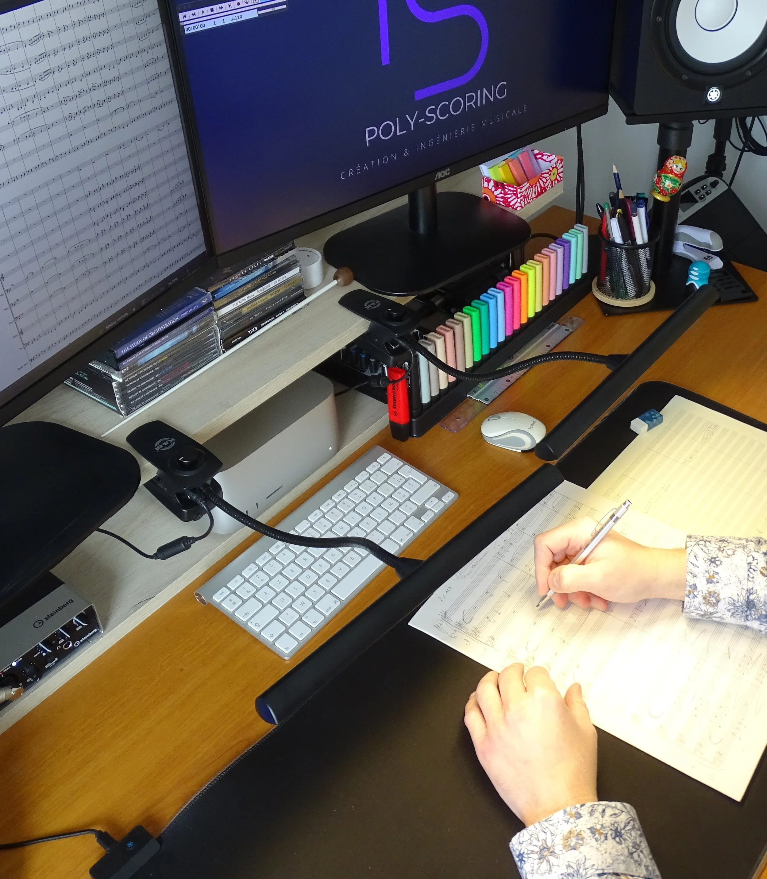 Bureau de travail avec deux écrans d'ordinateur, un clavier blanc, une souris, des instruments d'écriture, des cassettes musicales, une pince pour notes, et une main écrivant dans un carnet, avec stylos et pinceaux dans un porte-crayons. Deux haut-parleurs, une boîte à crayons, un carnet, et une main écrivant dans un cahier.