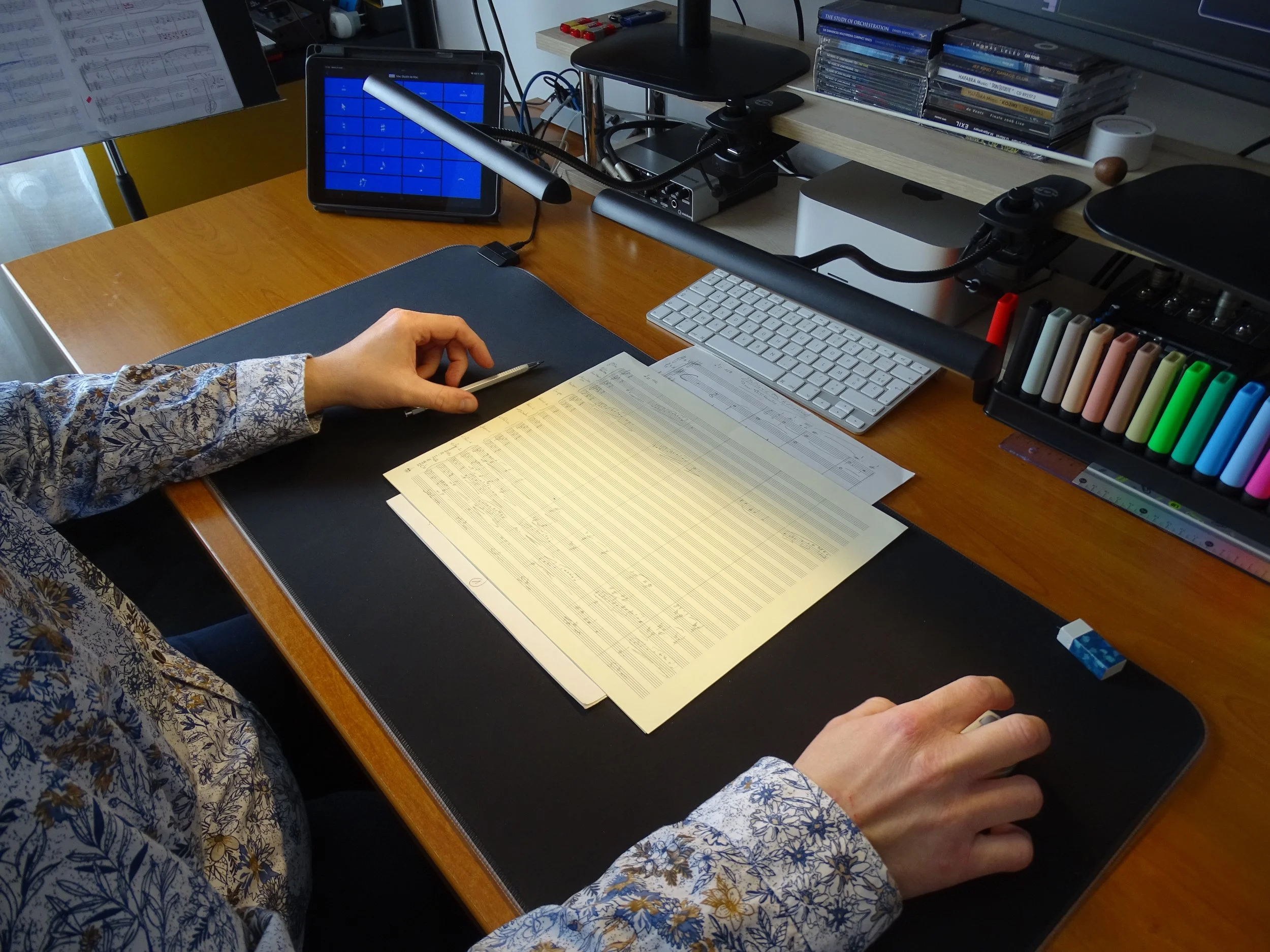 Un bureau avec une main d'une personne portant une chemise à fleurs blanches et bleues. Sur le bureau, il y a un clavier blanc, un écran avec une interface bleue, une souris, des documents, un stylo, et un établi avec des marqueurs de différentes couleurs. Au fond, il y a une étagère avec des CDs et des livres, et un support pour ordinateur.