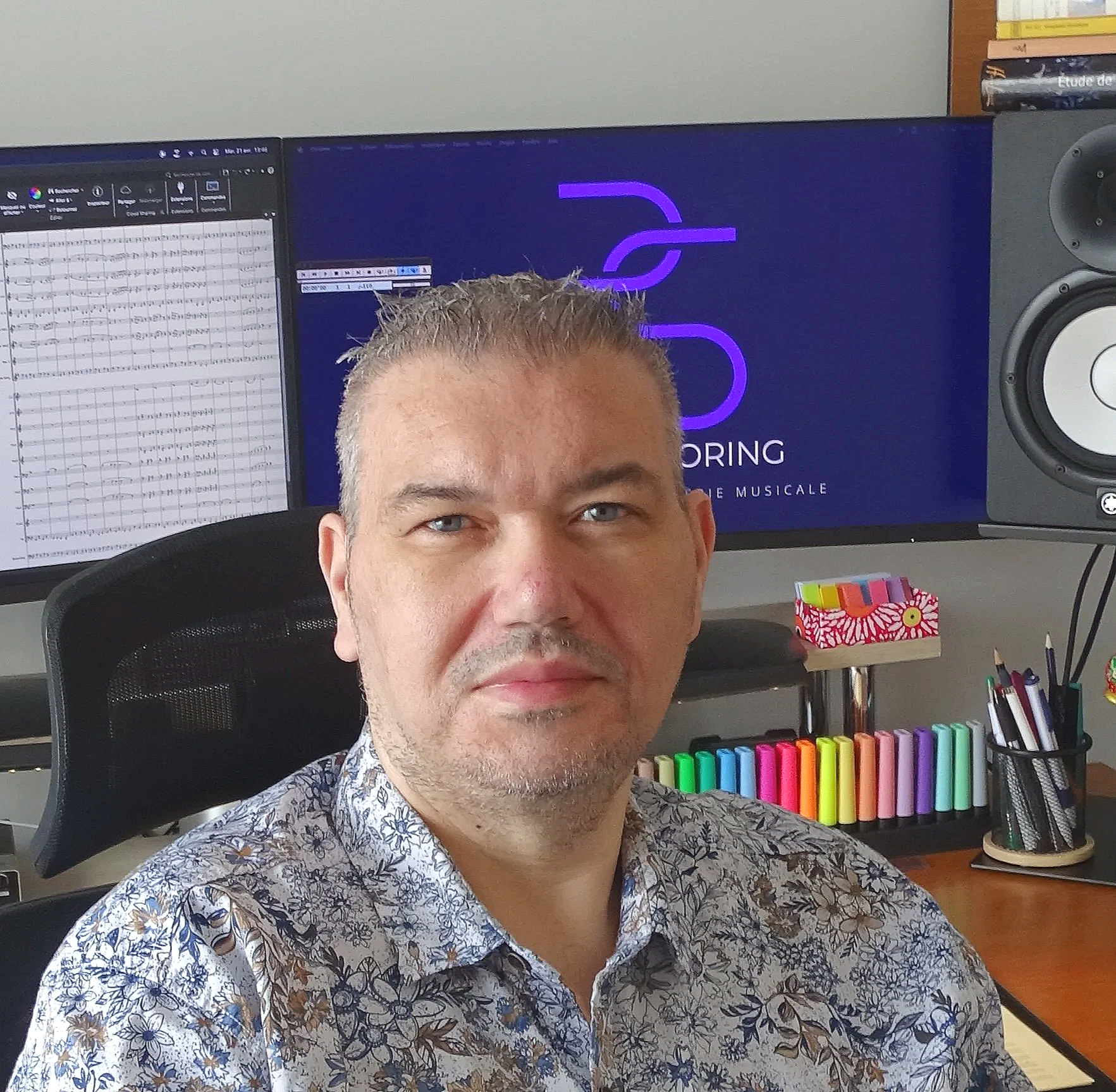 Un homme souriant avec des cheveux gris, portant une chemise à motifs floraux, assis devant un bureau avec des marqueurs de couleur, une enceinte, des livres et un ordinateur à deux écrans affichant des notes de musique et une présentation avec un logo violet.