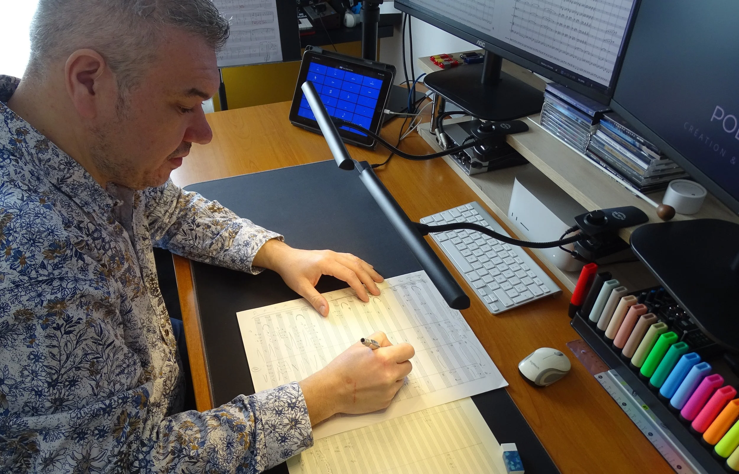 Homme avec des cheveux gris écrivant sur une partition musicale à un bureau avec deux écrans d'ordinateur, un clavier, une souris, un stylo, et plusieurs marqueurs colorés.